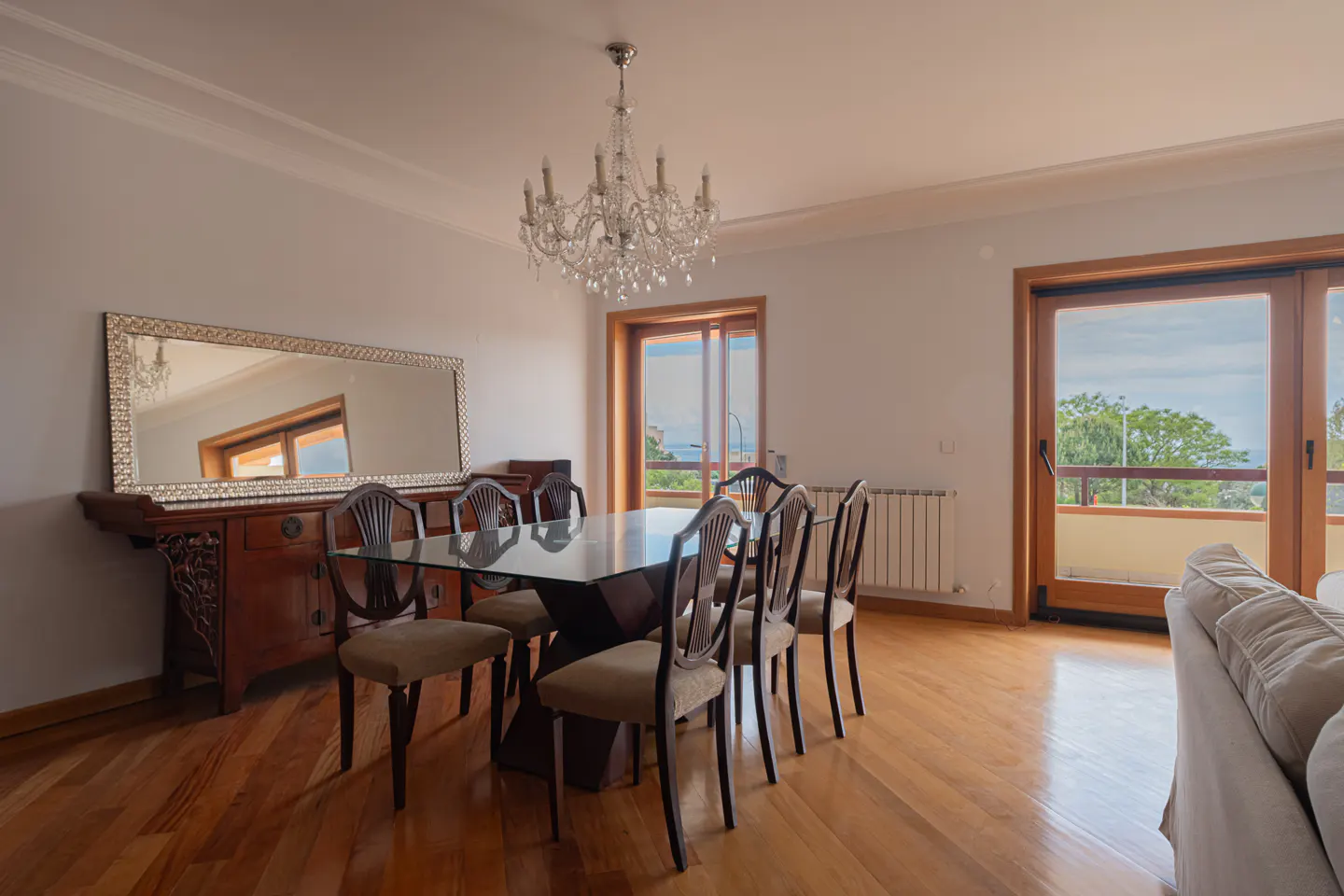 A dining room with a glass table, wooden chairs, and a crystal chandelier. A large mirror hangs above a wooden sideboard. Natural light streams in from the balcony.