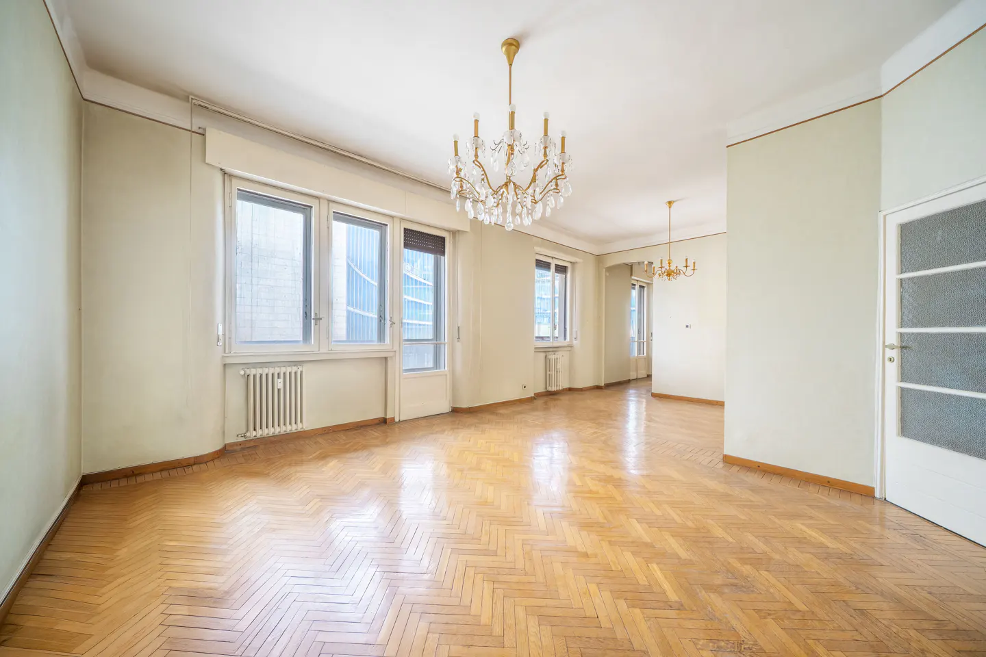 Empty room with herringbone wood floors, pale walls, and a crystal chandelier. Windows let in natural light.