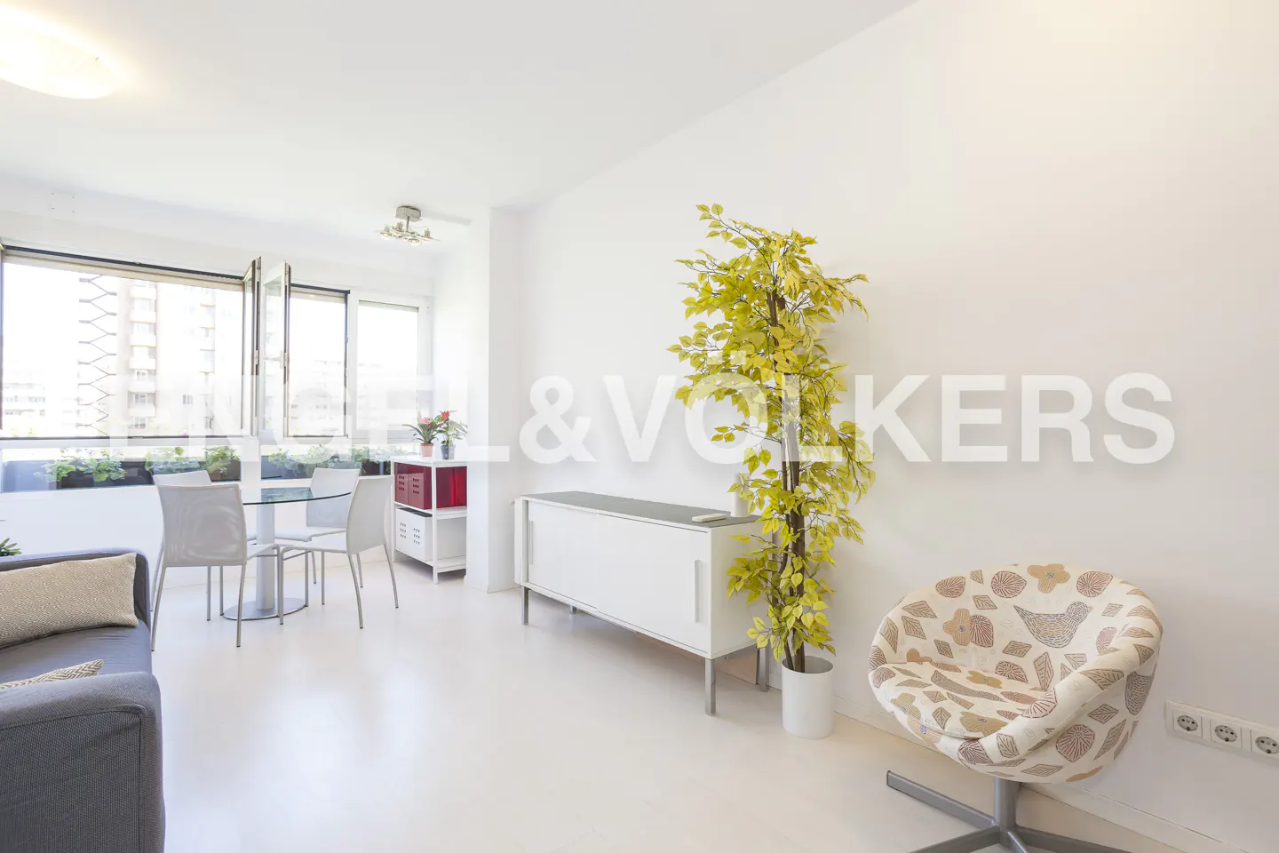 Bright living room with white walls, a white sideboard, a patterned armchair, and a dining area by a large window.