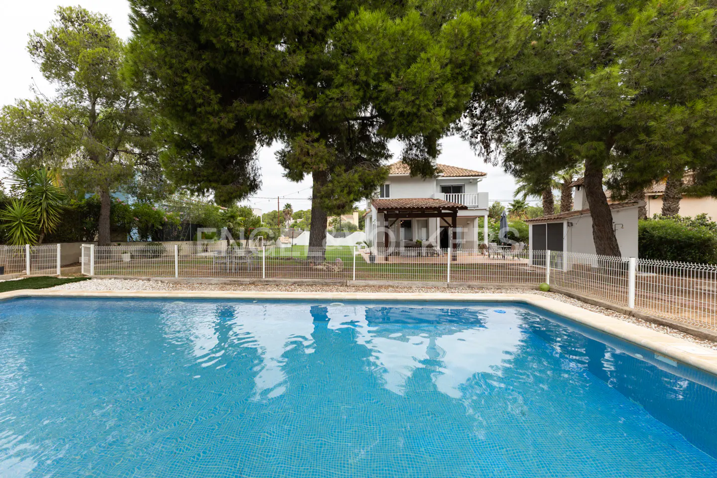 A bright blue tiled swimming pool in front of a white two-story house with trees.