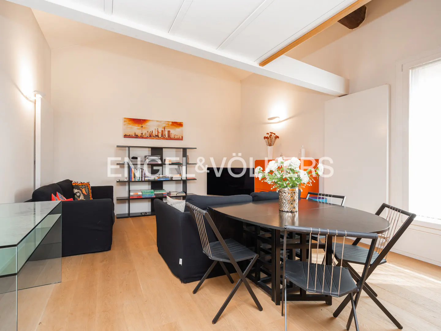 Bright living room with black sofas, a dark wood table with chairs, and light wood floors. Bookshelves and artwork add color.