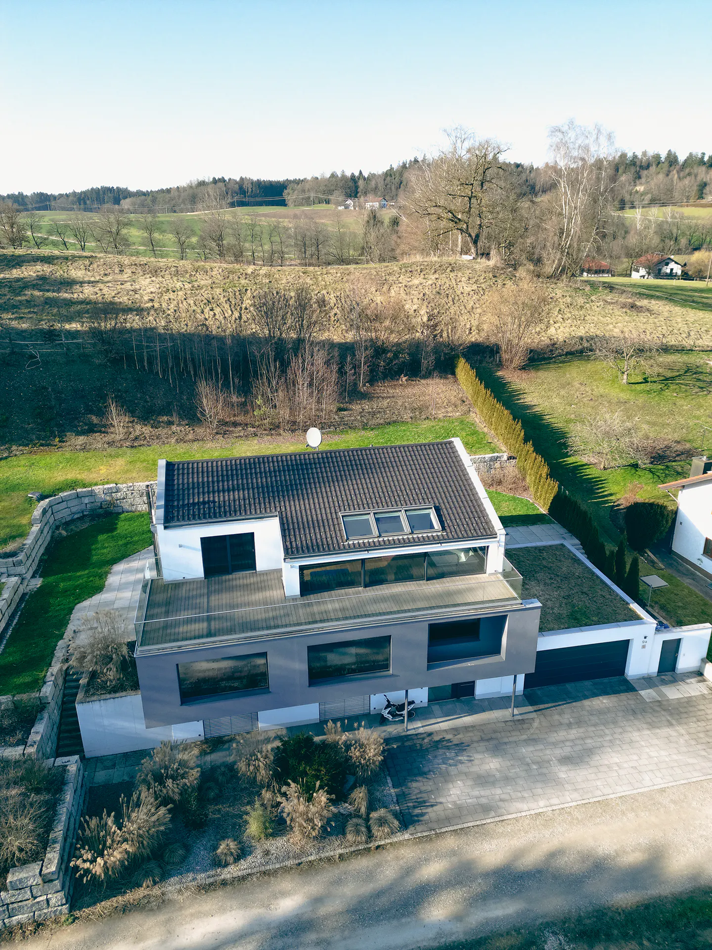 Aerial view of a modern, two-story house with a gray and white exterior, a dark roof with skylights, and a green roof over the garage. The house is surrounded by green lawns and trees.