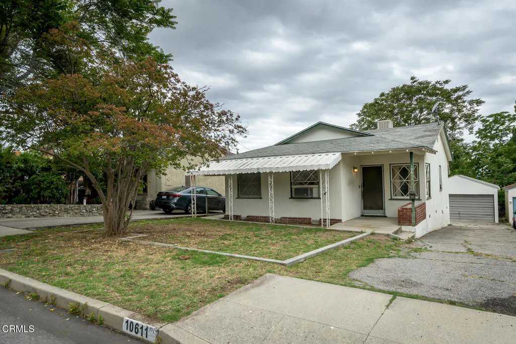 A white, single-story house with a white awning and green trim, a tree in the front yard, and a car parked in the driveway.