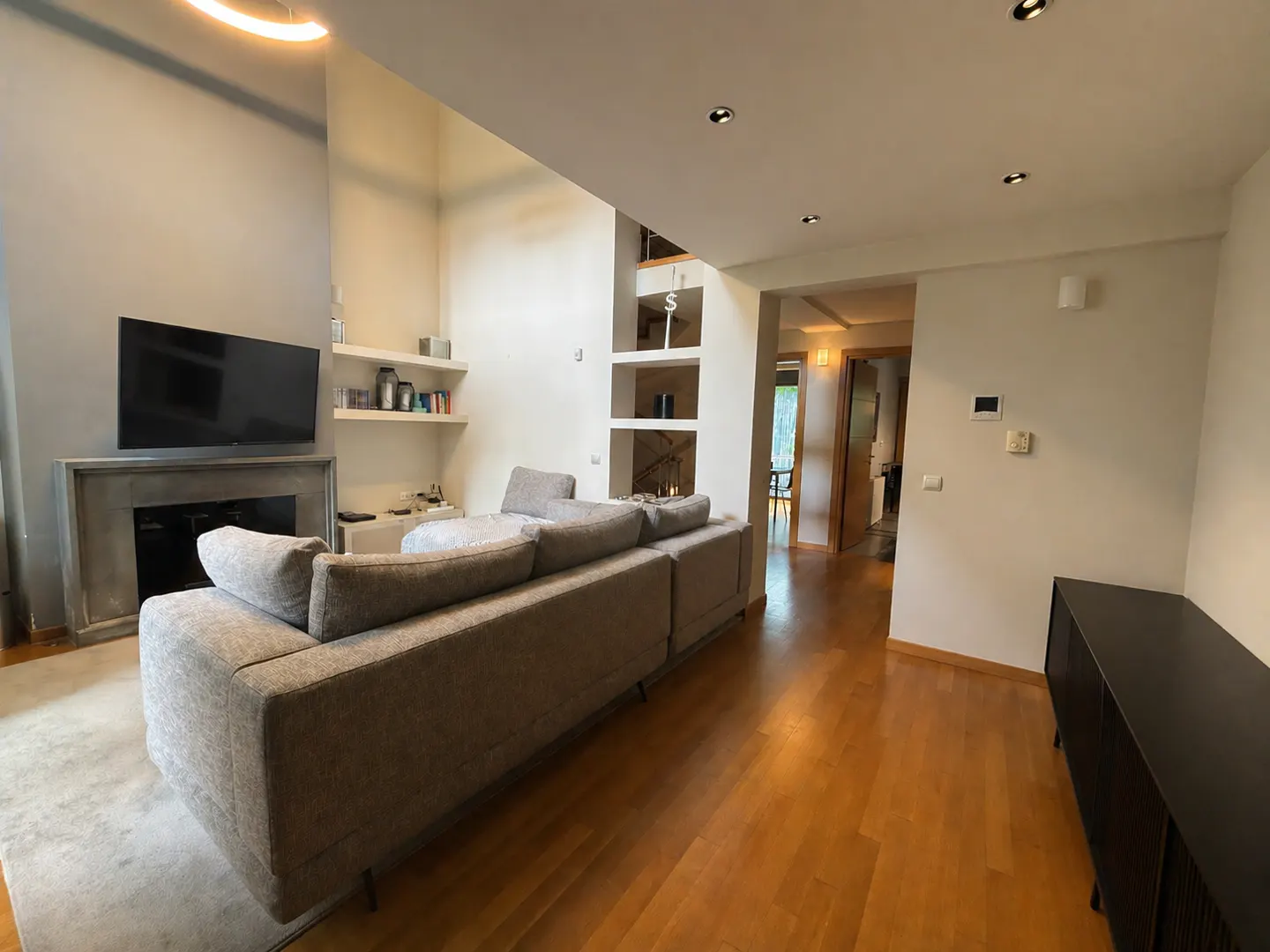 Living room with a gray sofa, TV over a fireplace, and hardwood floors. Shelves are built into the wall.