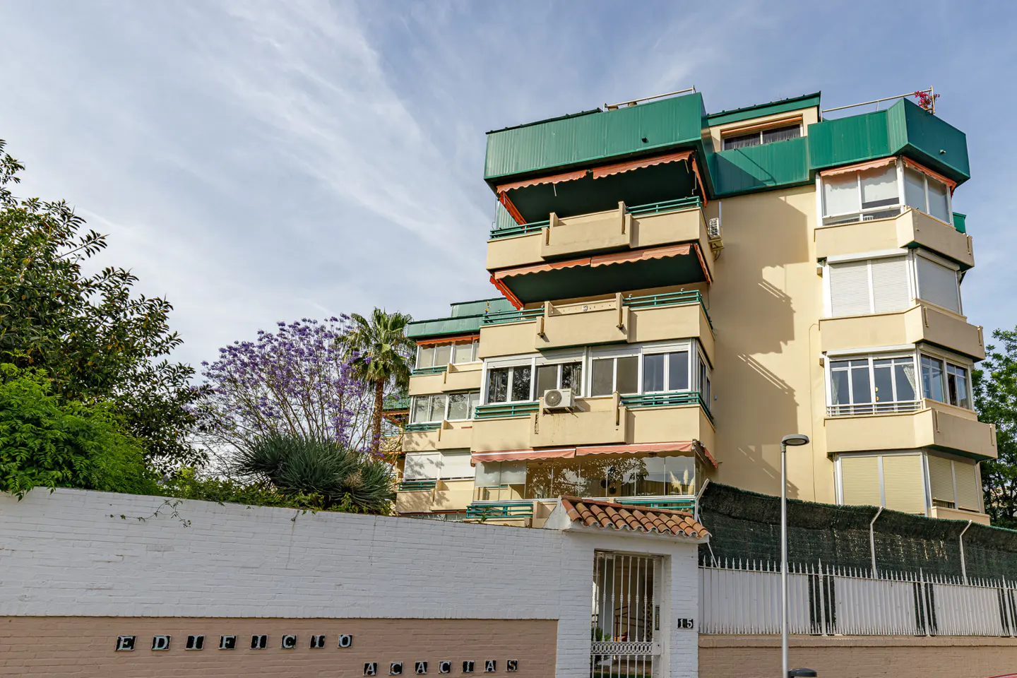 Exterior view of Edificio Acacias, a multi-story tan building with green awnings and balconies, behind a white brick wall and gate.