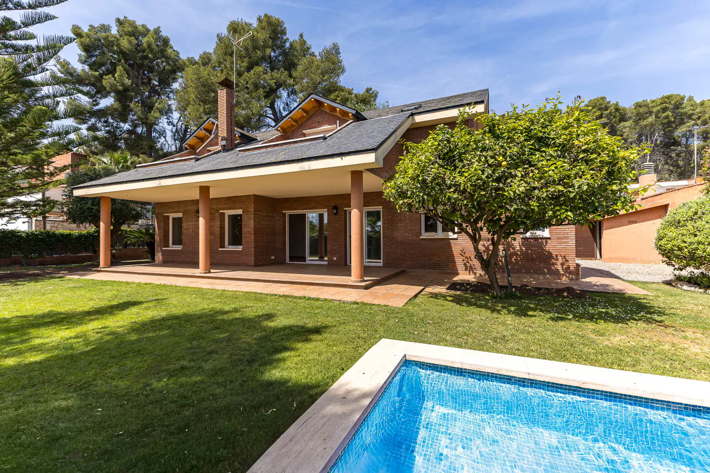 Brick house with a covered patio, green lawn, and a blue tiled pool under a sunny sky.