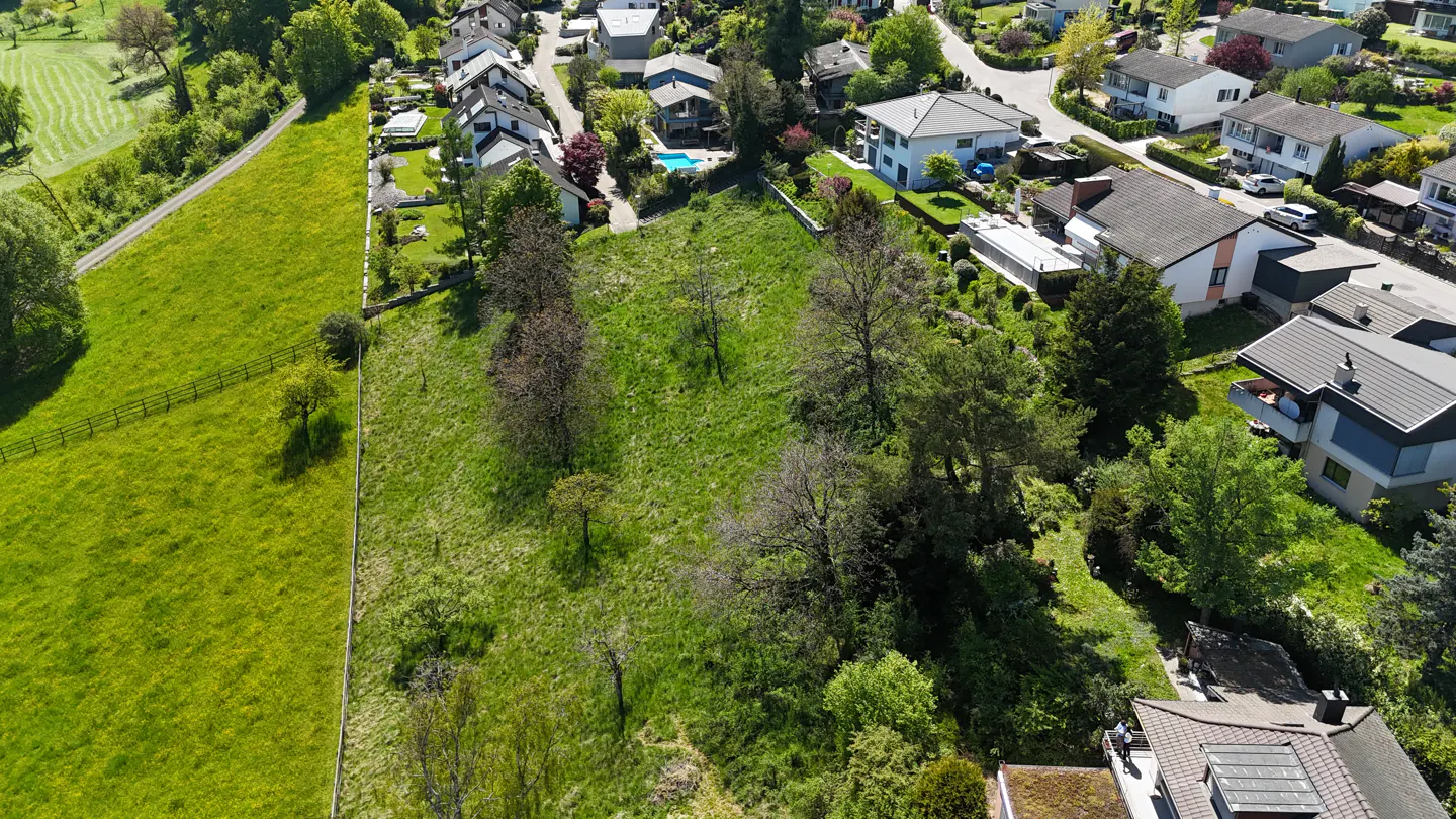 Aerial view of a grassy lot for sale, surrounded by houses and trees in a residential area. A green field borders the property.
