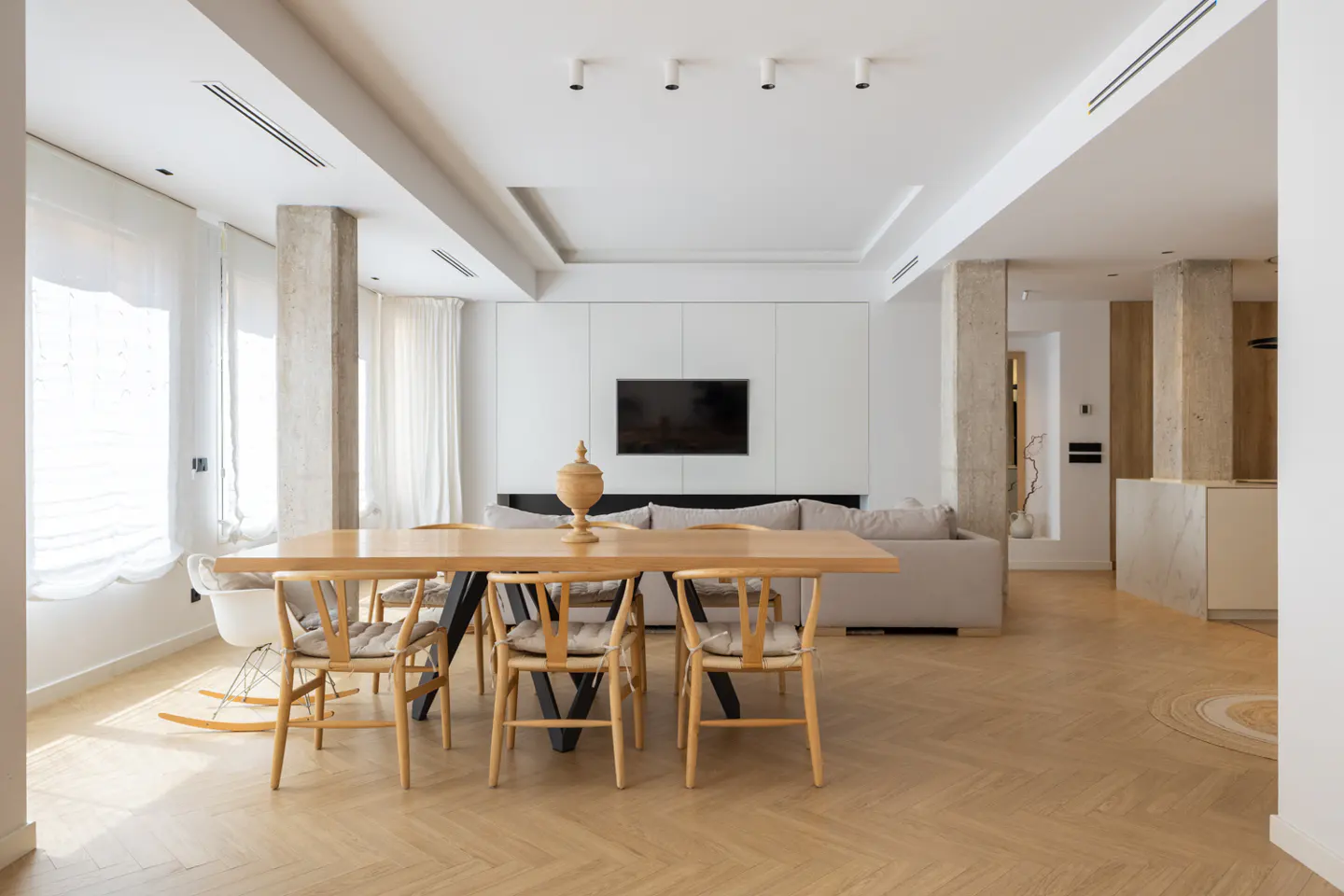Bright, modern dining room with a light wood table, chairs, and herringbone floors. A TV is mounted on a white wall above a gray sofa.