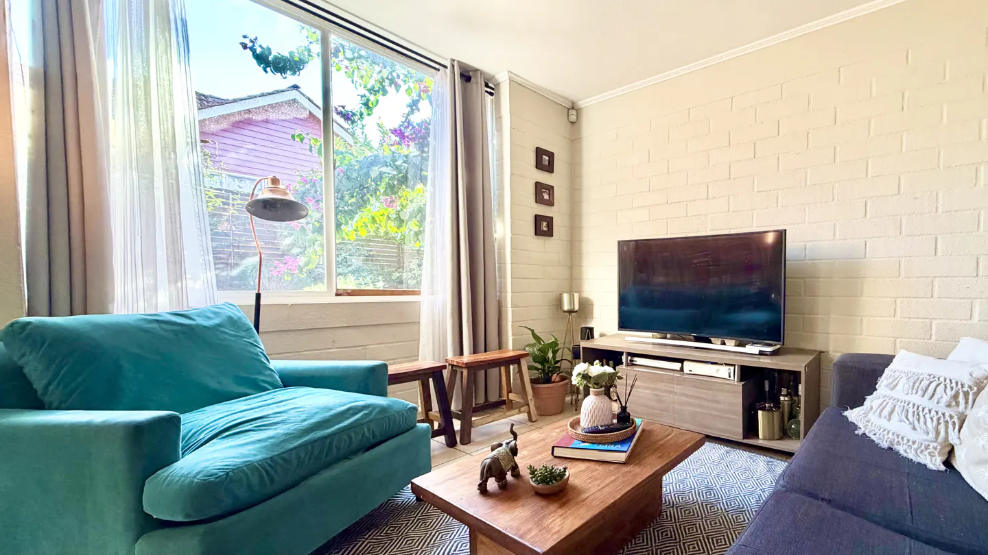 Living room with a teal armchair, wooden coffee table, TV, and a large window with garden view.