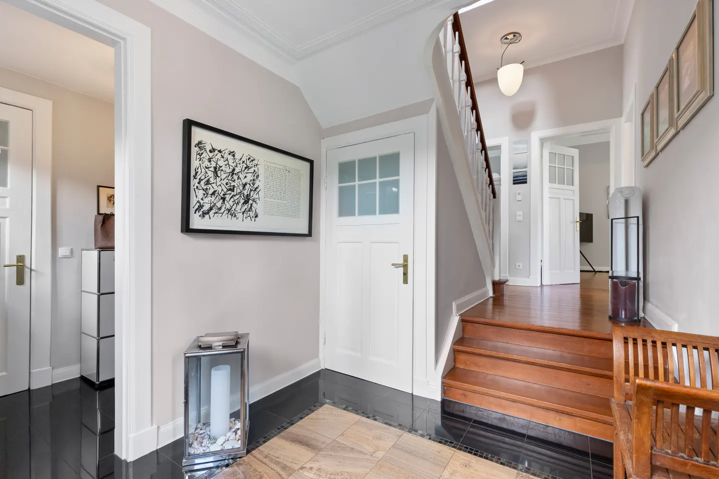 Hallway with black tile floor, wooden stairs, white walls, and doors. Artwork hangs on the wall. A silver lantern sits on the floor.