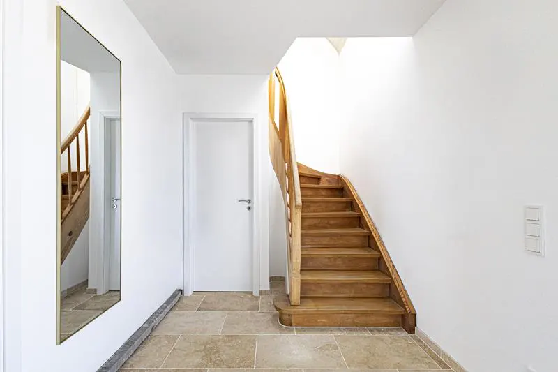 Bright hallway with stone tile floor, a white door, a wooden staircase, and a tall mirror reflecting the stairs.