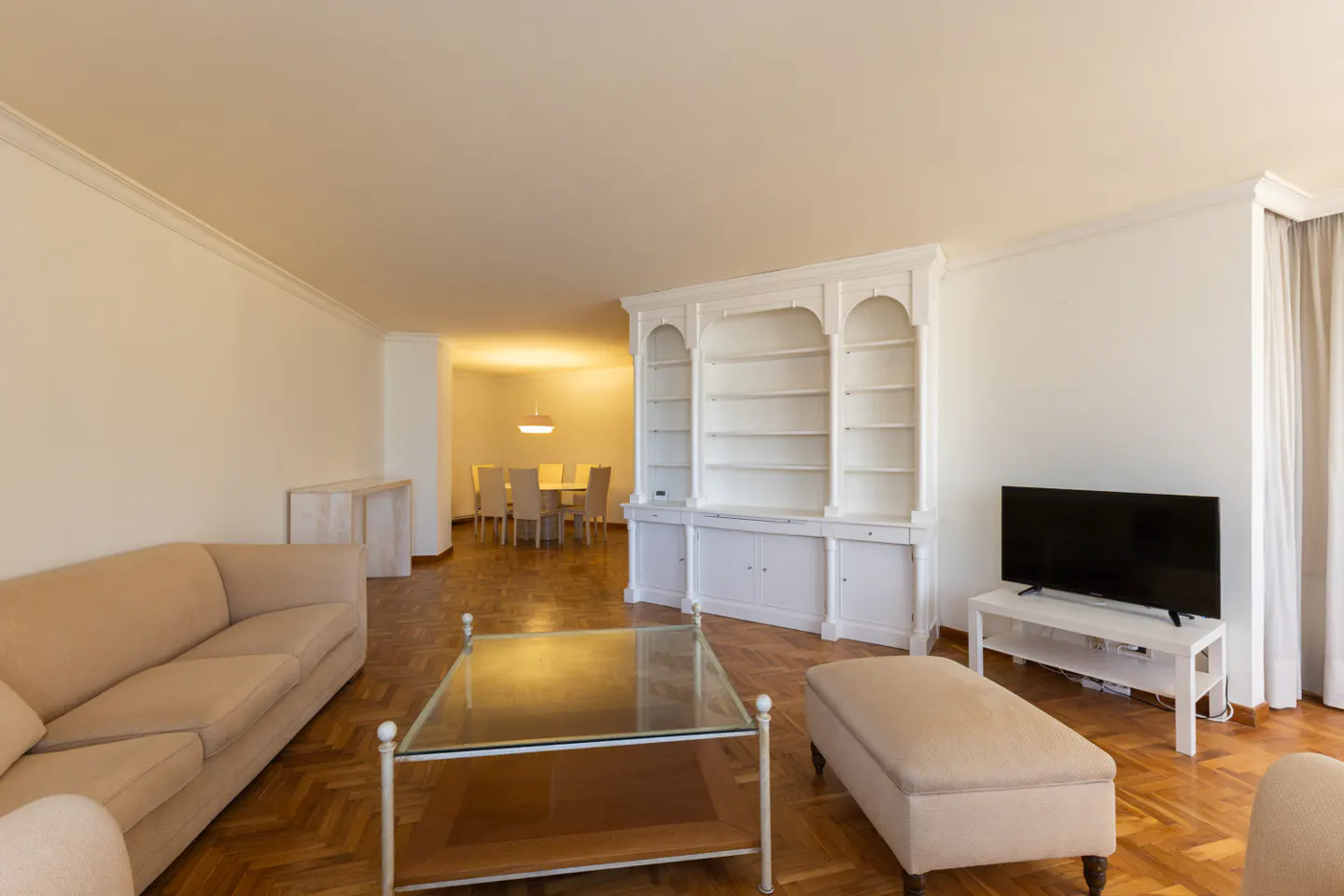 Living room with beige sofa, glass coffee table, white bookcase, and TV. Dining room visible in background.