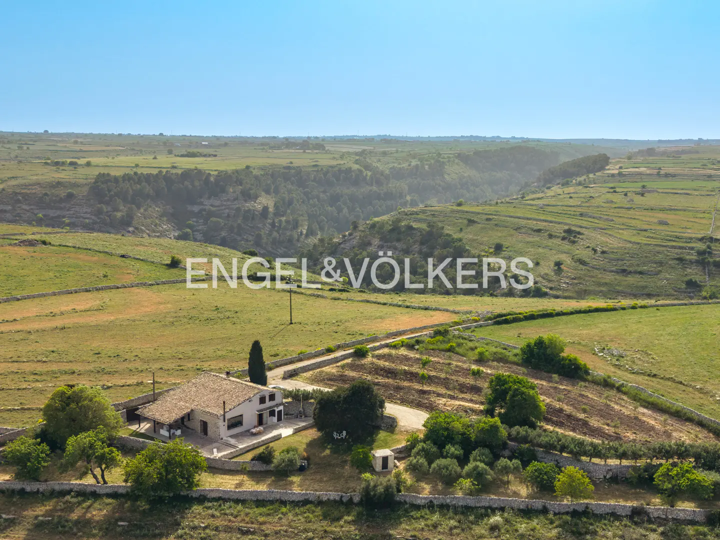 Aerial view of a stone farmhouse with a tiled roof surrounded by green fields, trees, and stone walls in a rural landscape.