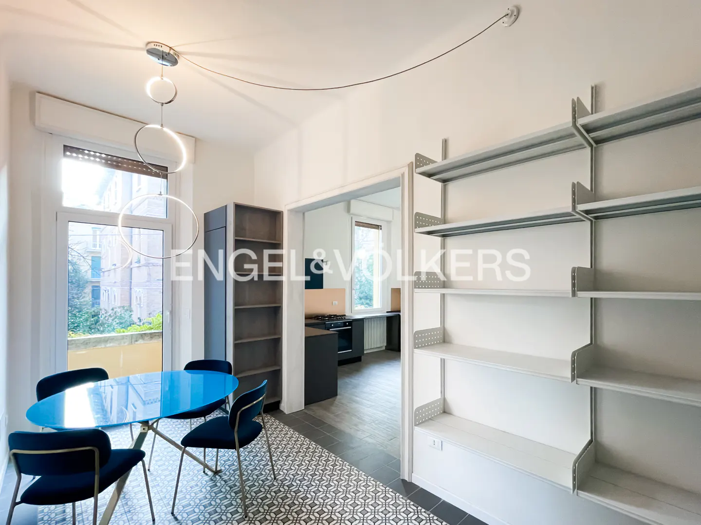 Bright apartment interior with a blue glass table, navy chairs, and white shelving. A kitchen is visible through an open doorway.