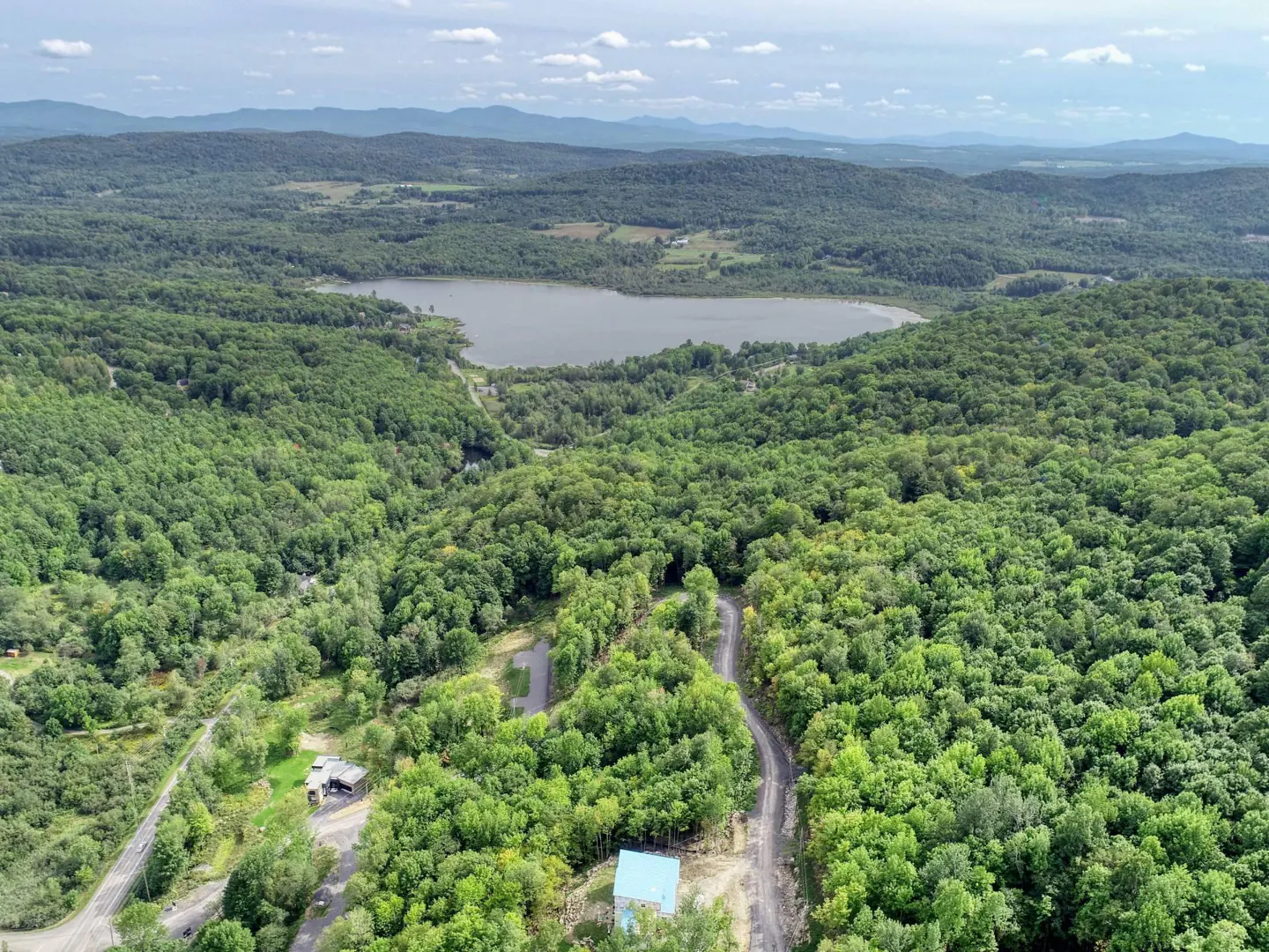 Aerial view of a lake surrounded by green forest and mountains under a blue sky with white clouds. A dirt road leads to a house with a blue roof.