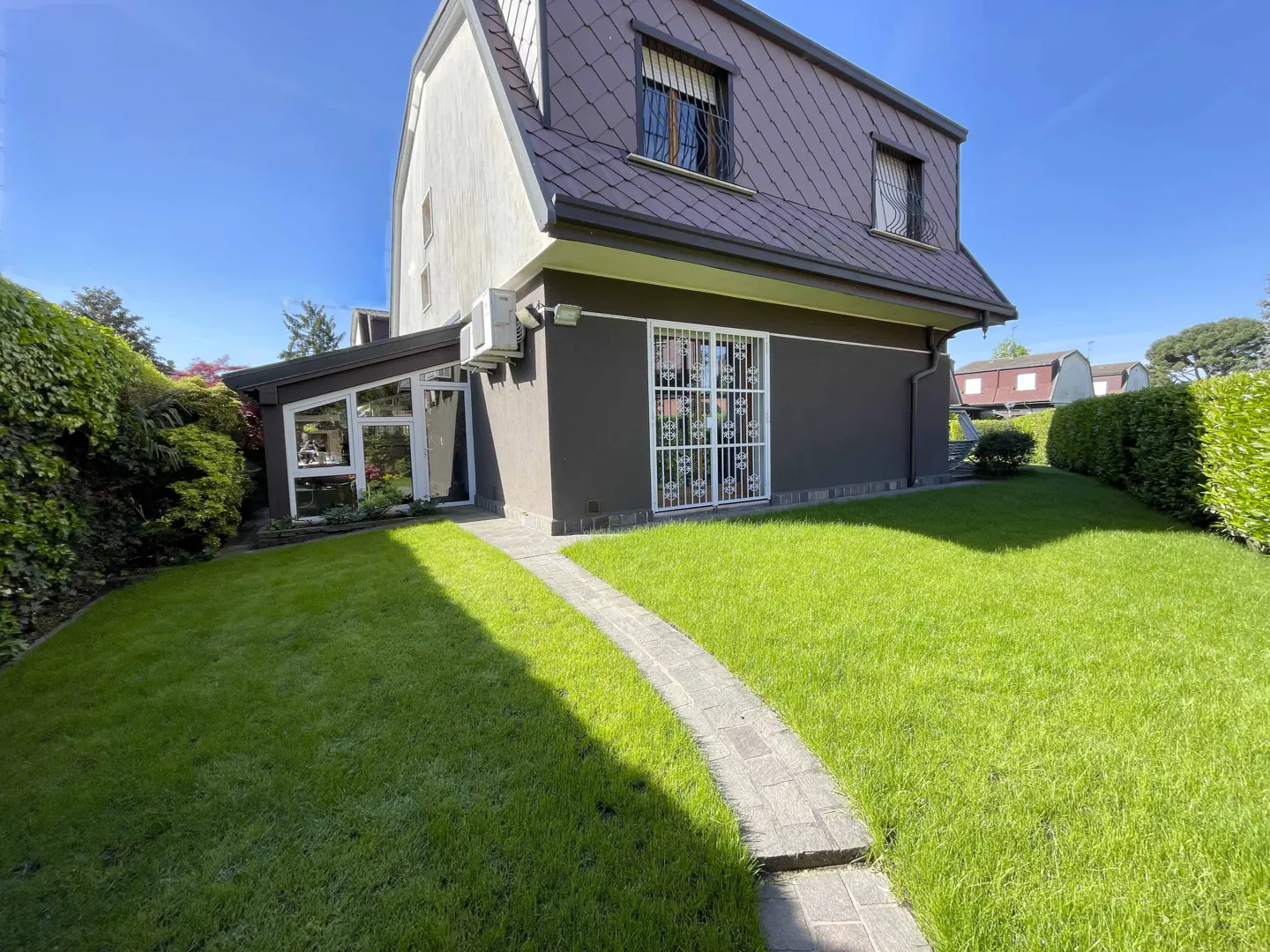 Two-story house with brown roof, white and brown walls, and green lawn. A stone path leads to a white-grilled door.