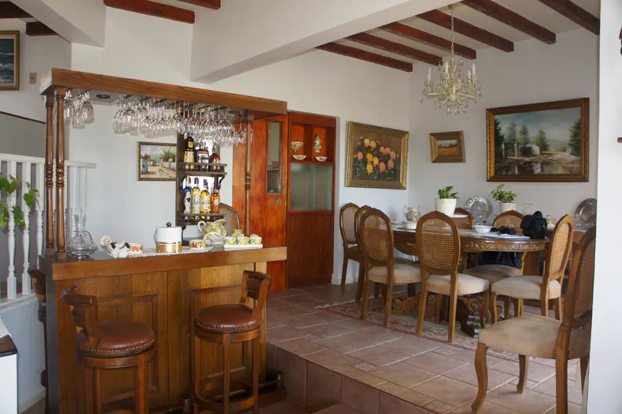 Interior view of a dining room with a wooden bar, stools, and a dining table with chairs. Paintings adorn the walls.