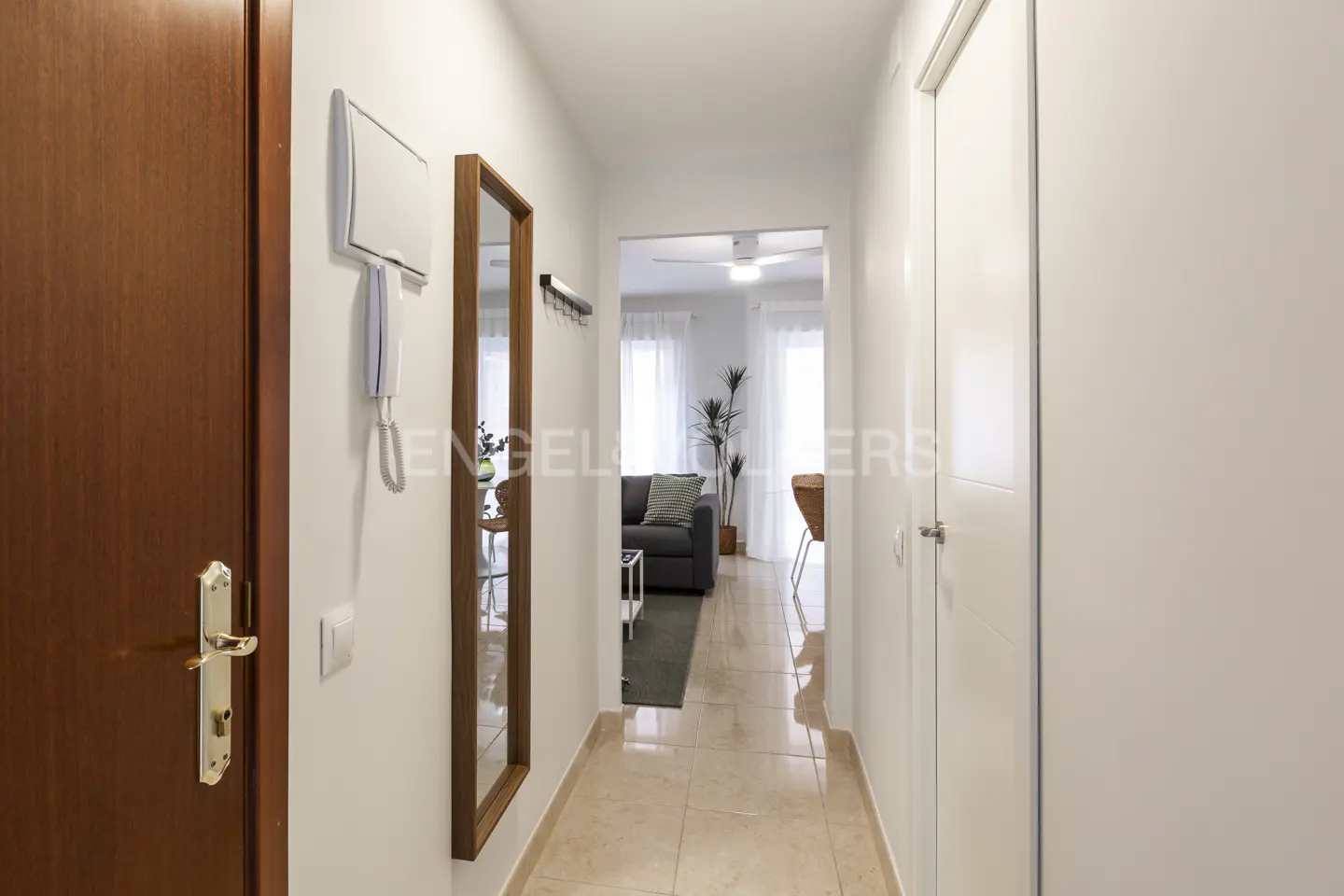 Hallway with a brown door, intercom, mirror, and coat rack. Living room with a gray sofa and plant visible in the background.