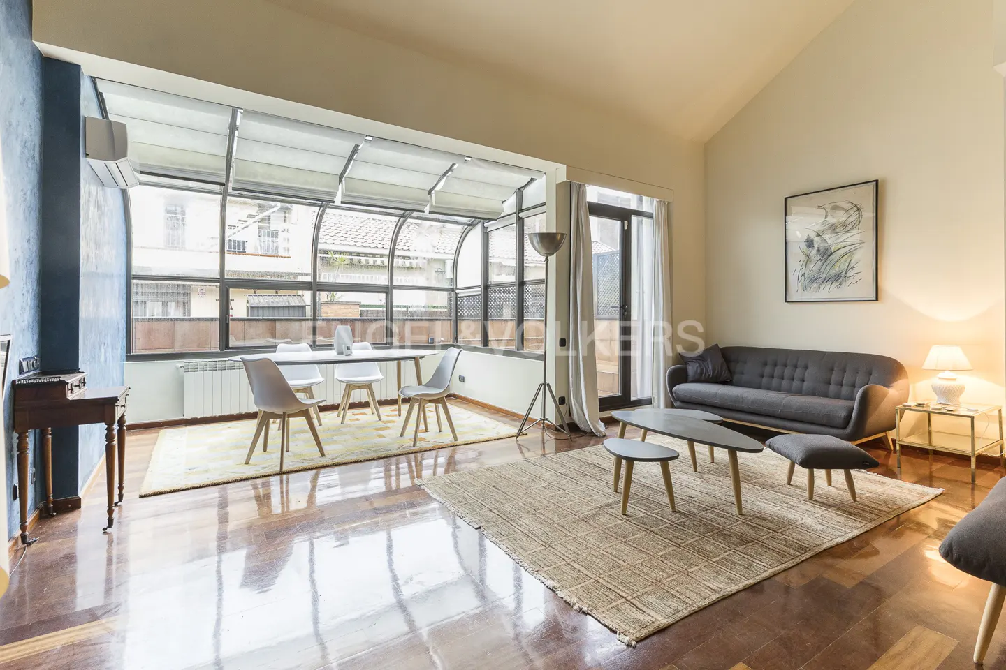 Bright living room with hardwood floors, a gray sofa, and a dining table by a large window. A tan rug adds warmth to the space.