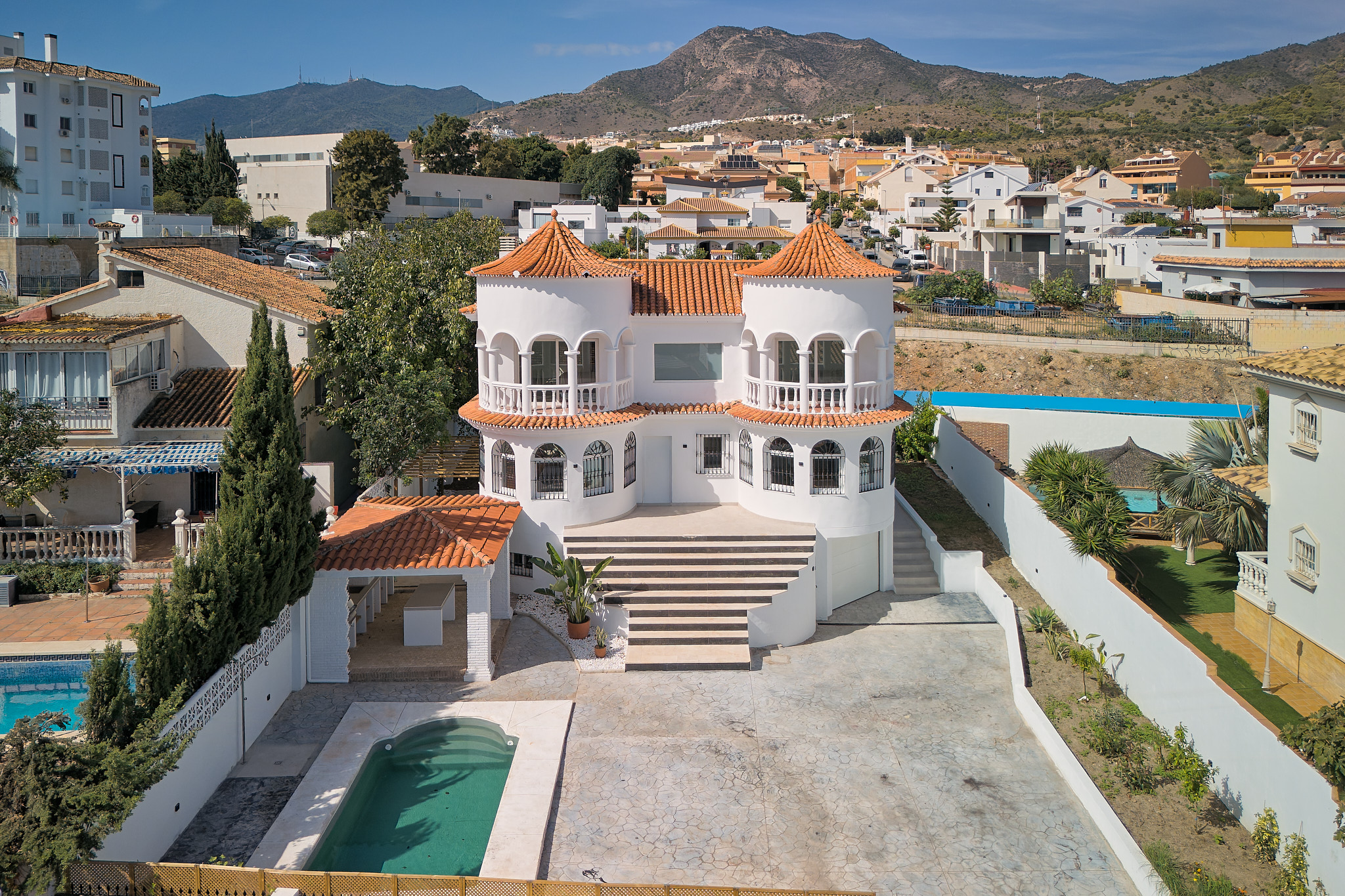 White two-story villa with twin towers, orange tile roofs, a swimming pool, and a large front patio in a sunny neighborhood. [AI-generated text]