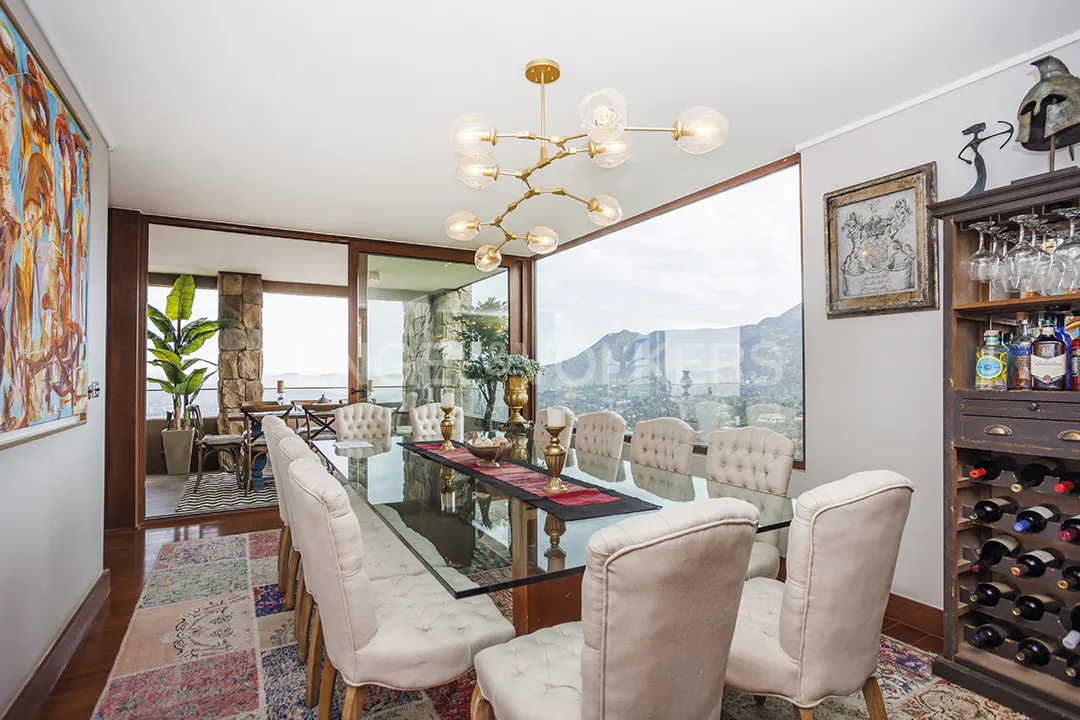 A dining room with a glass table, upholstered chairs, and a gold chandelier. A large window shows a mountain view.