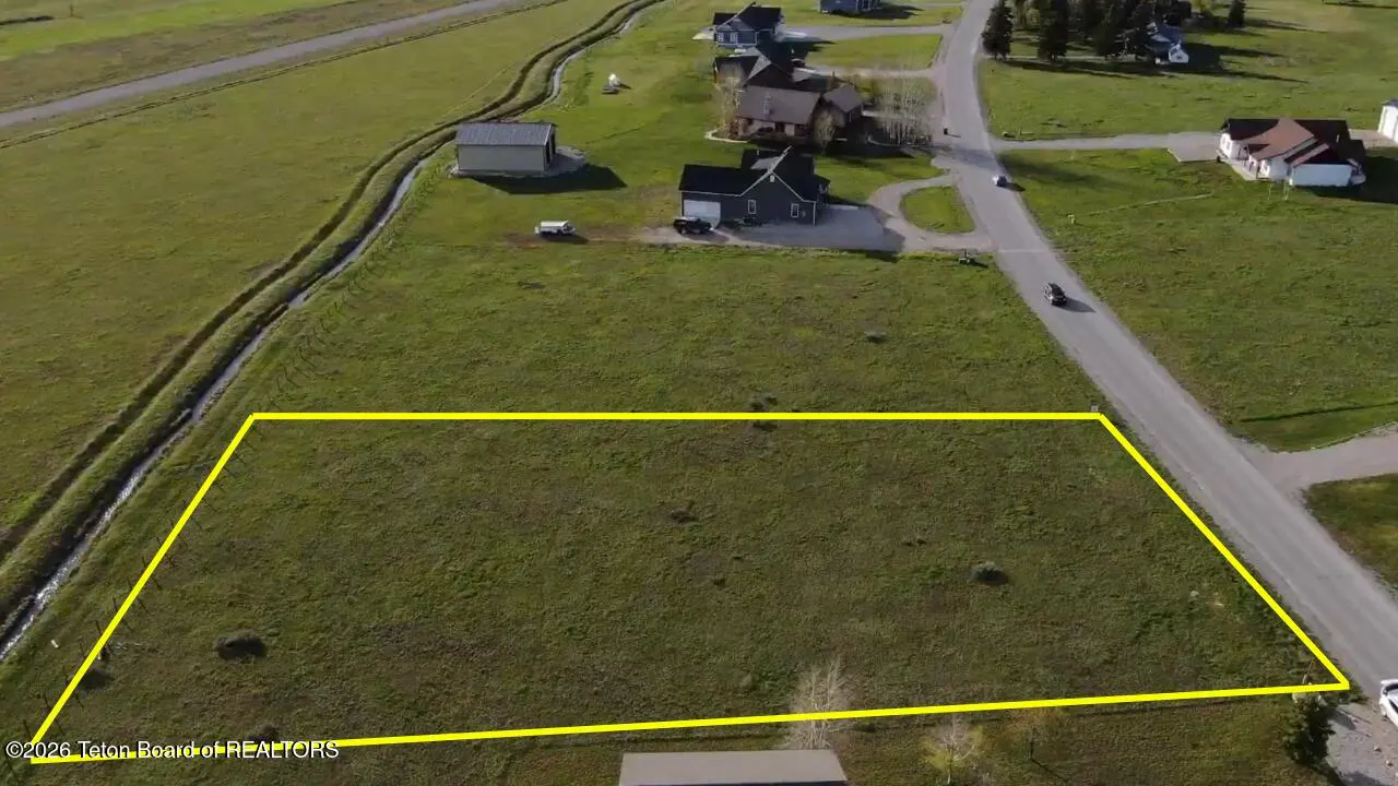 Aerial view of a vacant, grassy rectangular lot outlined in yellow, situated in a residential neighborhood near a road.