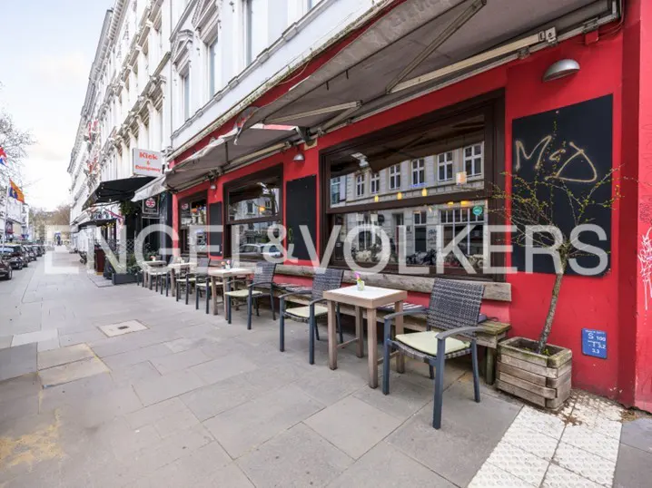 Outdoor cafe with red walls, awning, and tables with chairs on a stone sidewalk. Building reflections in the windows.