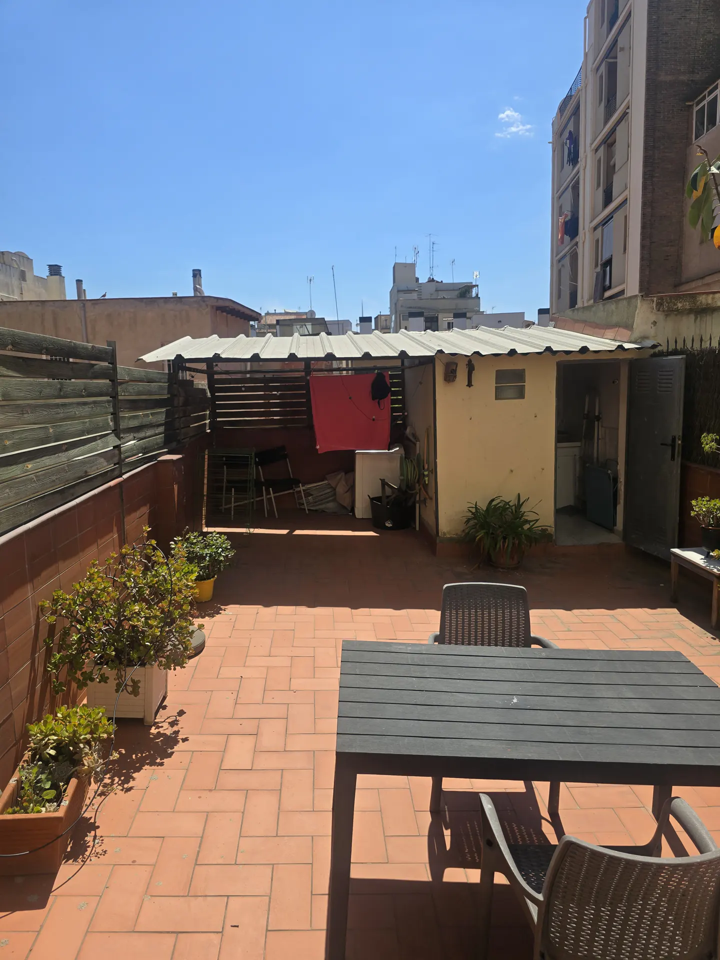Rooftop patio with brick tiles, a dark table and chairs. A shed and fence line the back, with buildings in the background under a blue sky.