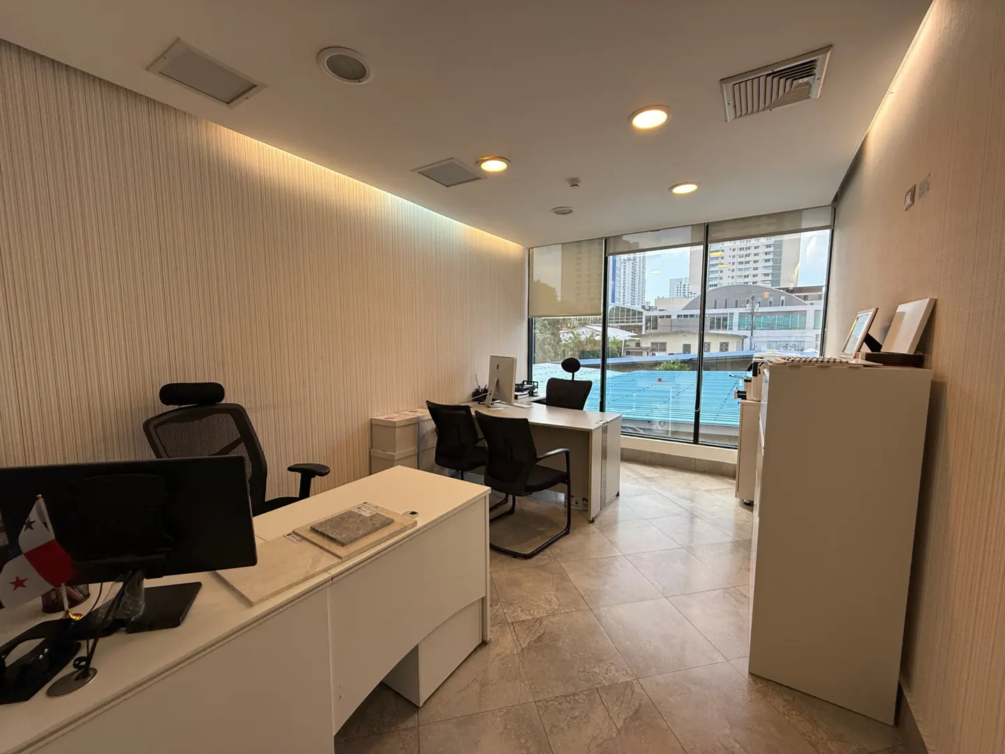 Bright office space with two desks, black chairs, and a large window overlooking a city. A Panama flag sits on the front desk.