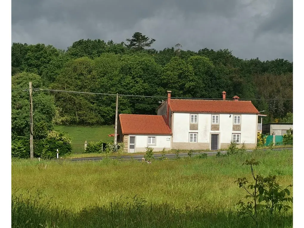 A two-story white house with a red tile roof sits in a green field near a forest under a cloudy sky.