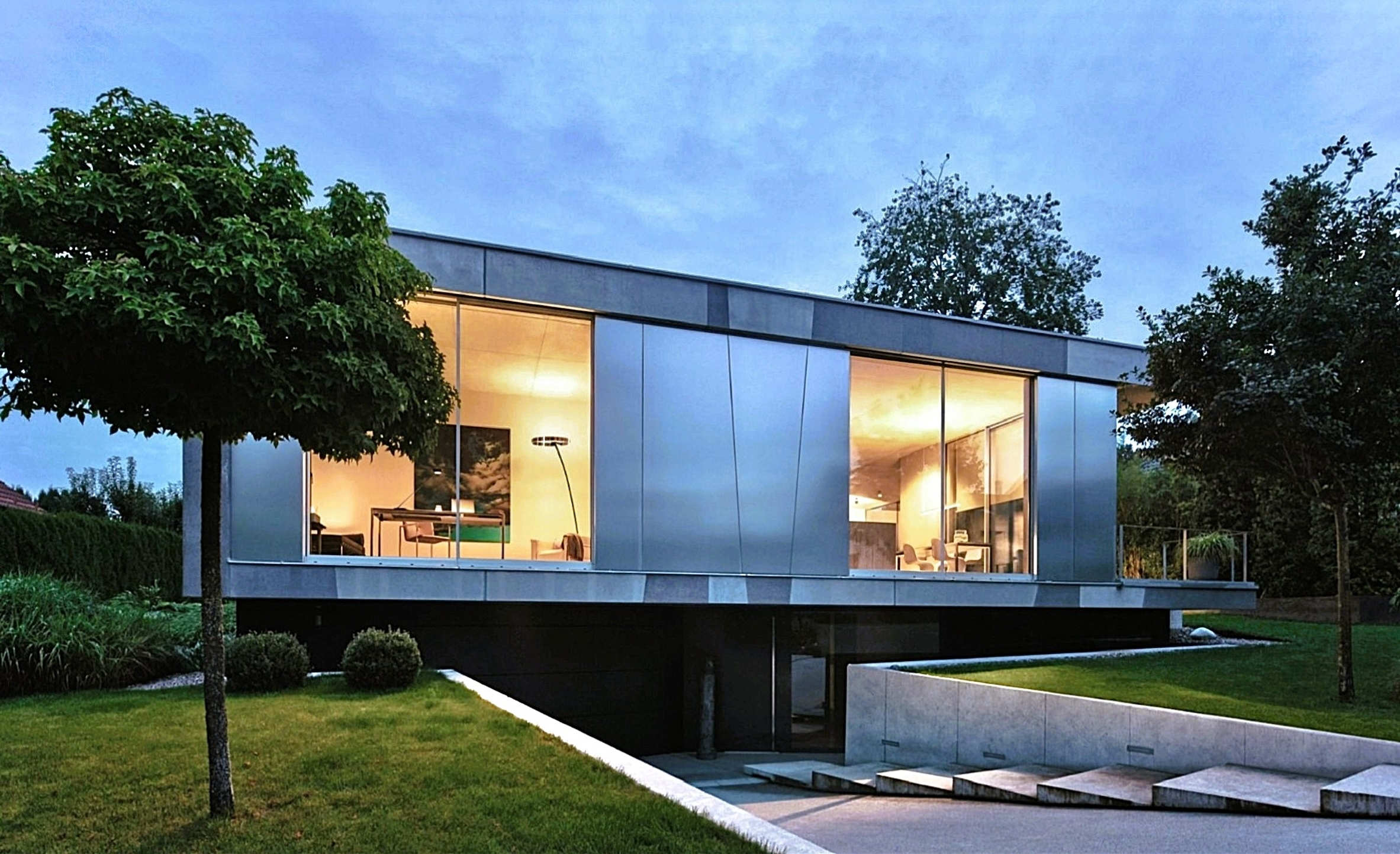 Modern two-story home with metal siding, large illuminated windows, and a landscaped yard at twilight. [AI-generated text]