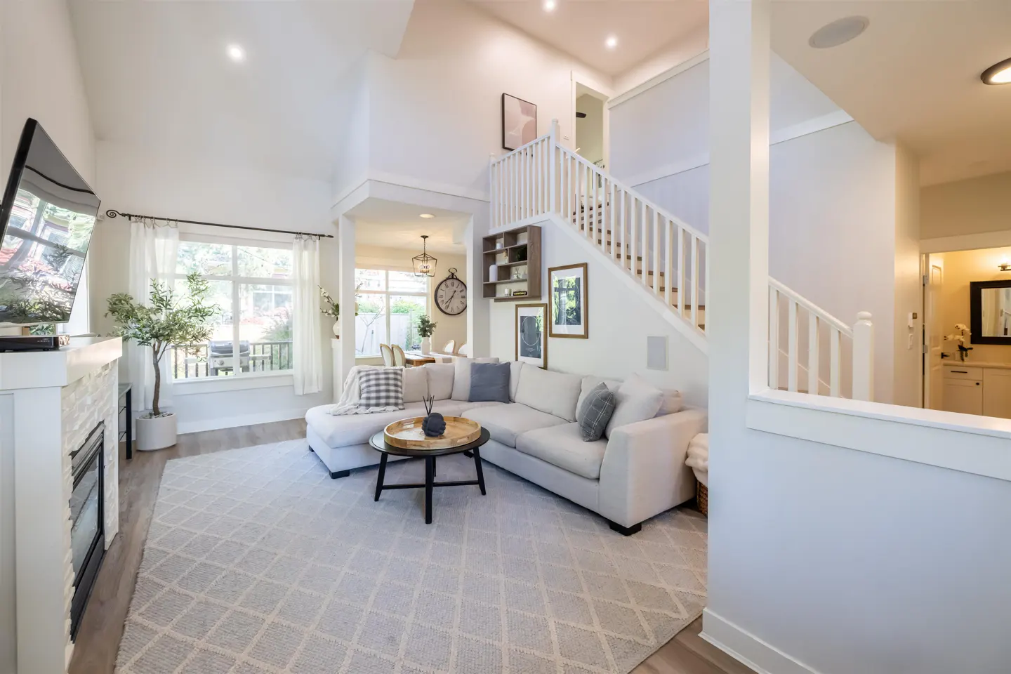 Bright, open-concept living room with white walls, a sectional sofa, and a staircase. A fireplace and large window add to the inviting atmosphere.