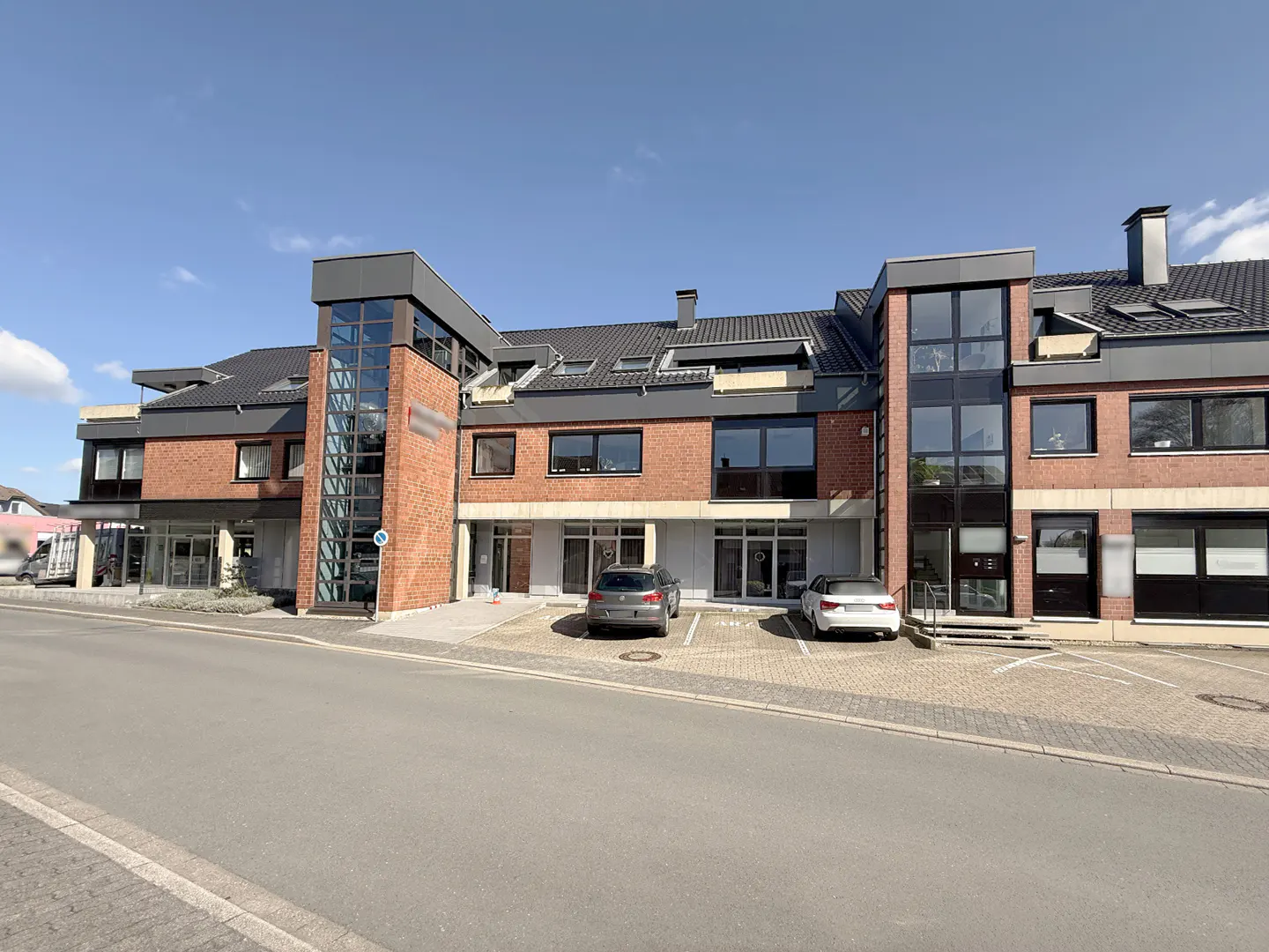 Two-story brick commercial building with black trim and glass tower accents. Two cars are parked in front on a sunny day.