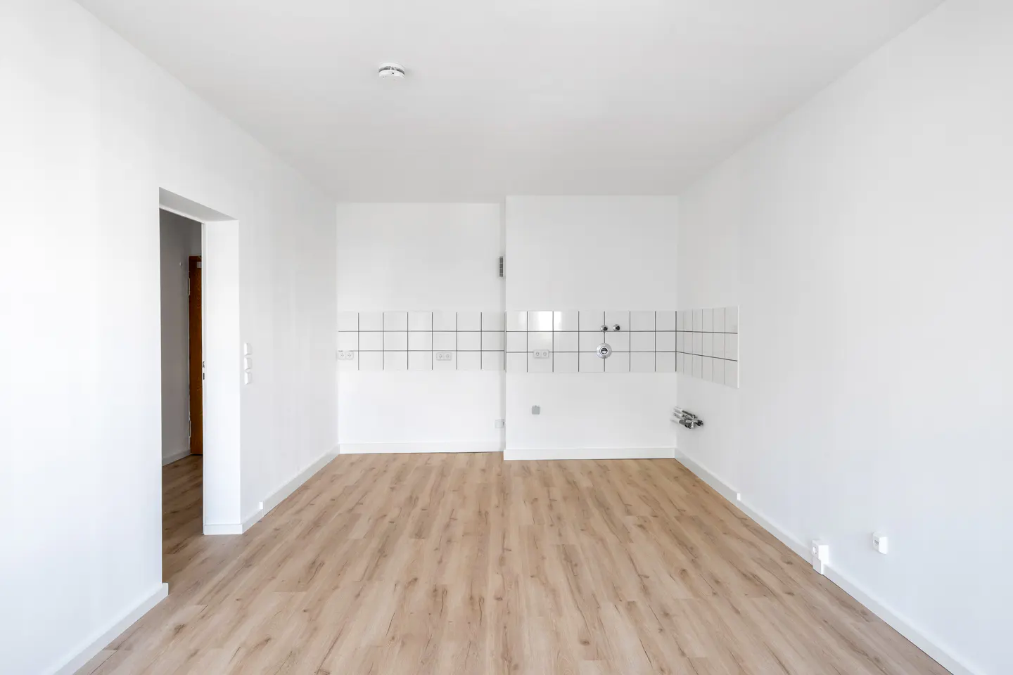 Empty room with white walls, light wood-look flooring, and a white tiled backsplash area for a kitchen installation.