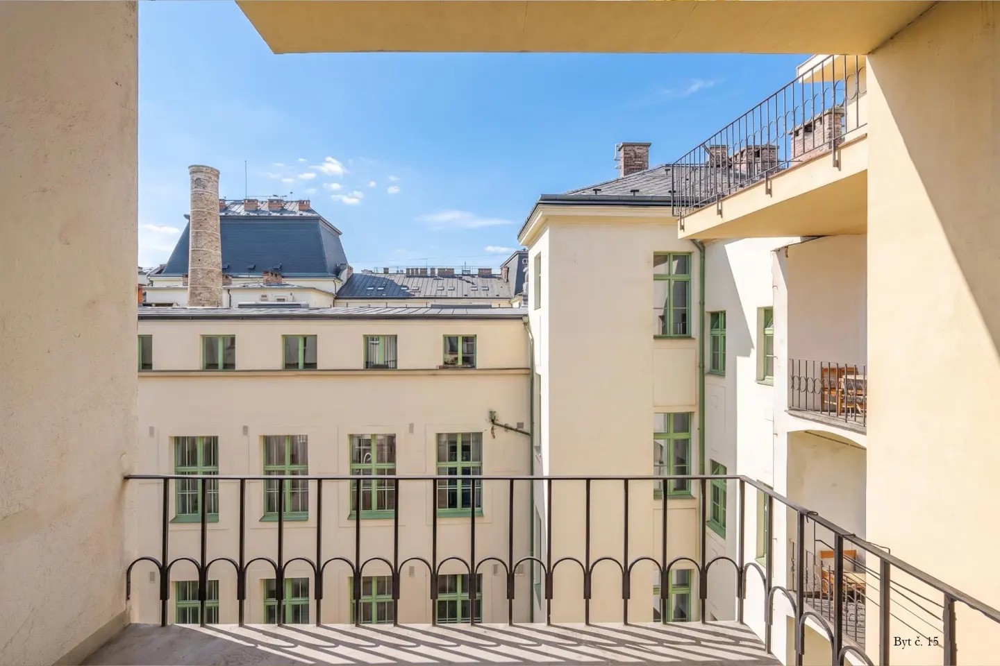 View from a balcony overlooking a sunny courtyard surrounded by cream-colored apartment buildings with green-framed windows.