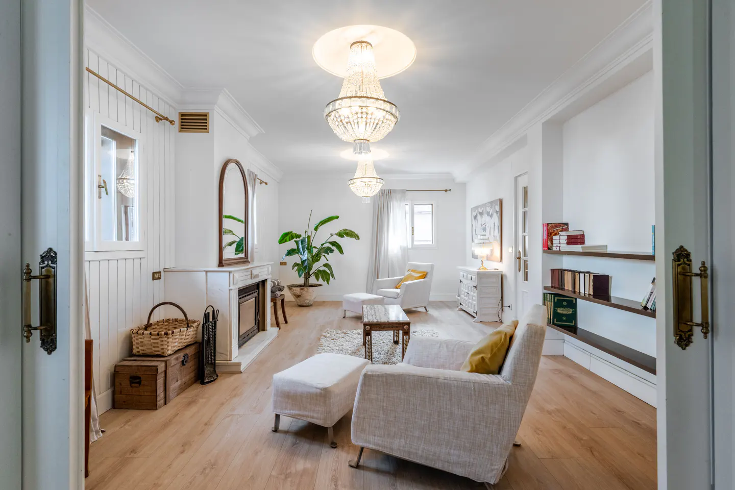 Bright living room with hardwood floors, white walls, and a fireplace. Crystal chandeliers hang from the ceiling. Armchairs and a plant add warmth.
