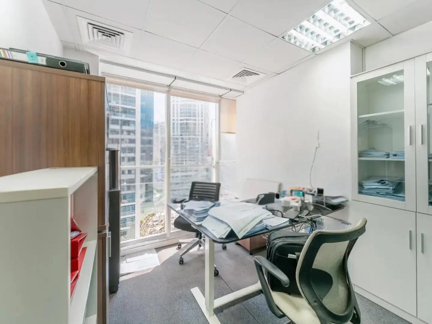 Bright office space with a desk, rolling chairs, and a large window overlooking a city skyline. White walls and cabinets.