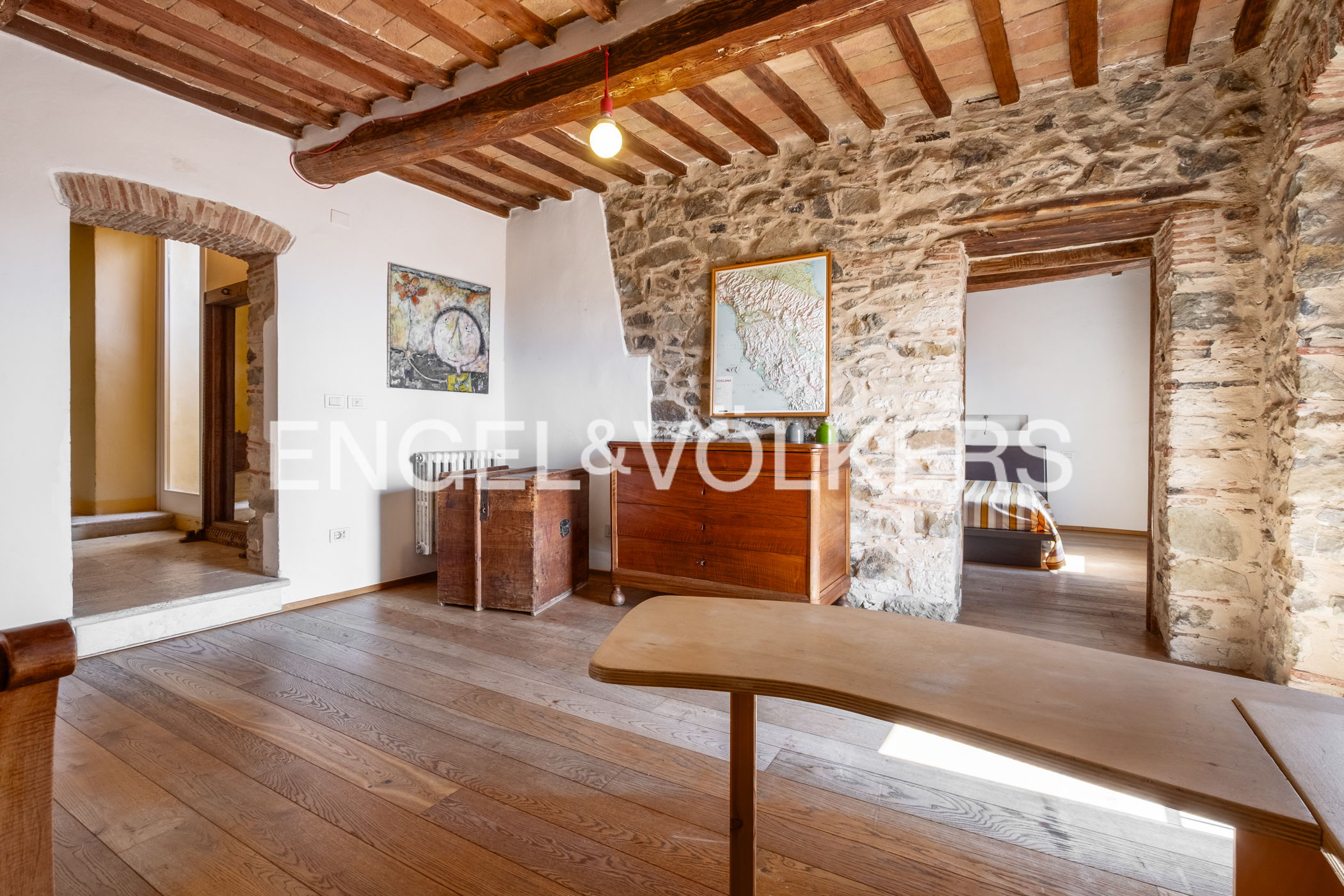 Interior view of a rustic room with stone walls, wooden beams, and hardwood floors. A wooden dresser and map adorn the wall. A table is in the foreground.