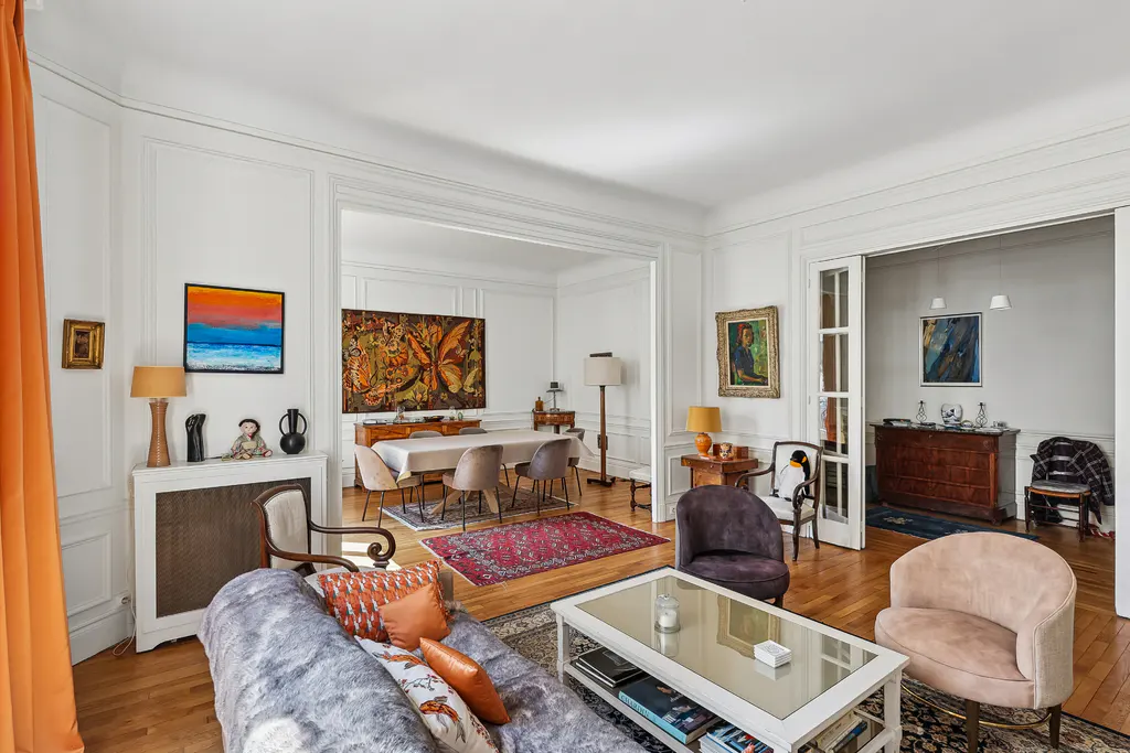 Bright, airy living room with hardwood floors, white walls, and colorful art. A dining area is visible through an archway.