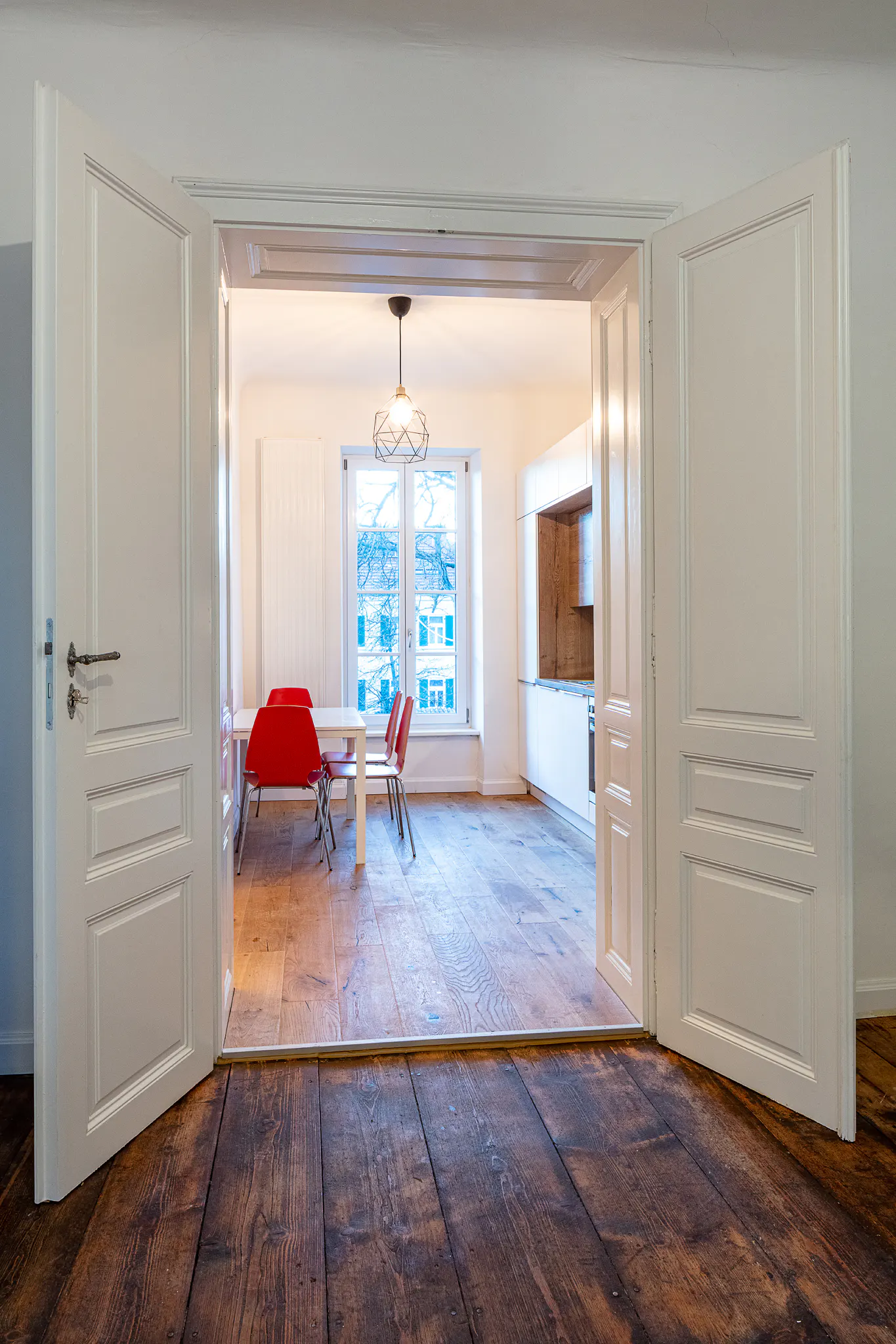 View through open white double doors into a bright dining room with red chairs, a white table, and a kitchen area.