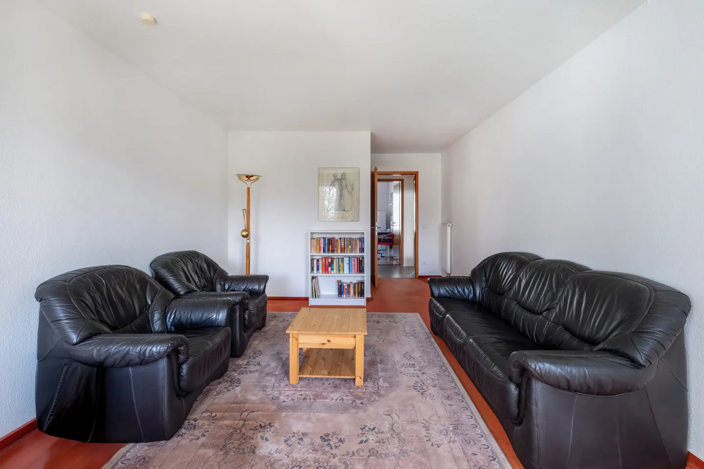 Living room with black leather sofa and armchairs, a wooden coffee table, and a bookshelf against white walls.