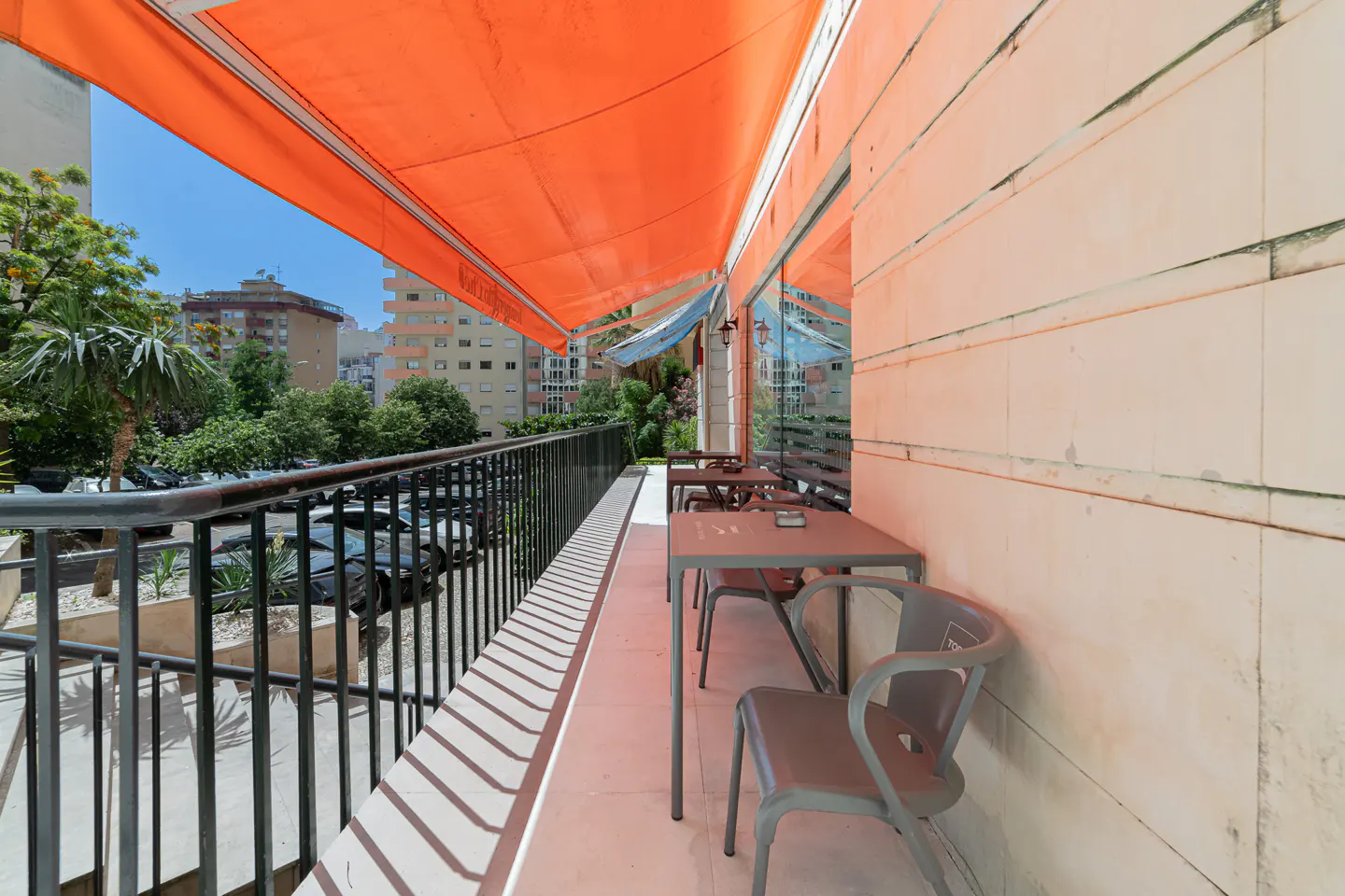 Outdoor cafe seating with metal tables and chairs under an orange awning. A black metal railing overlooks trees and buildings.