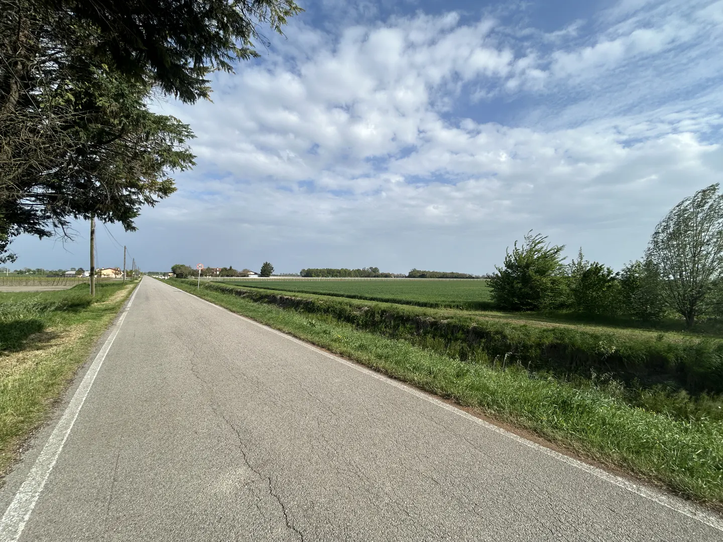 A long, gray asphalt road stretches into the distance, bordered by green fields and trees under a cloudy sky.