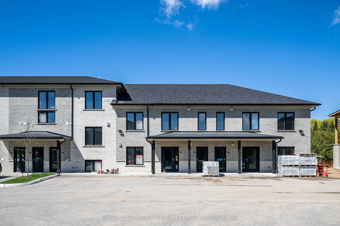 Exterior of a two-story, light gray brick townhouse with black trim under a clear blue sky. Pallets of materials sit in front.
