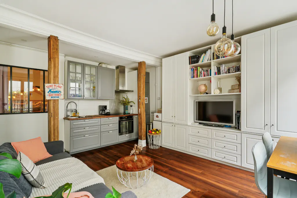 Modern open-plan living area with a grey kitchen, wooden support beams, a grey sofa, and a large white built-in bookshelf.