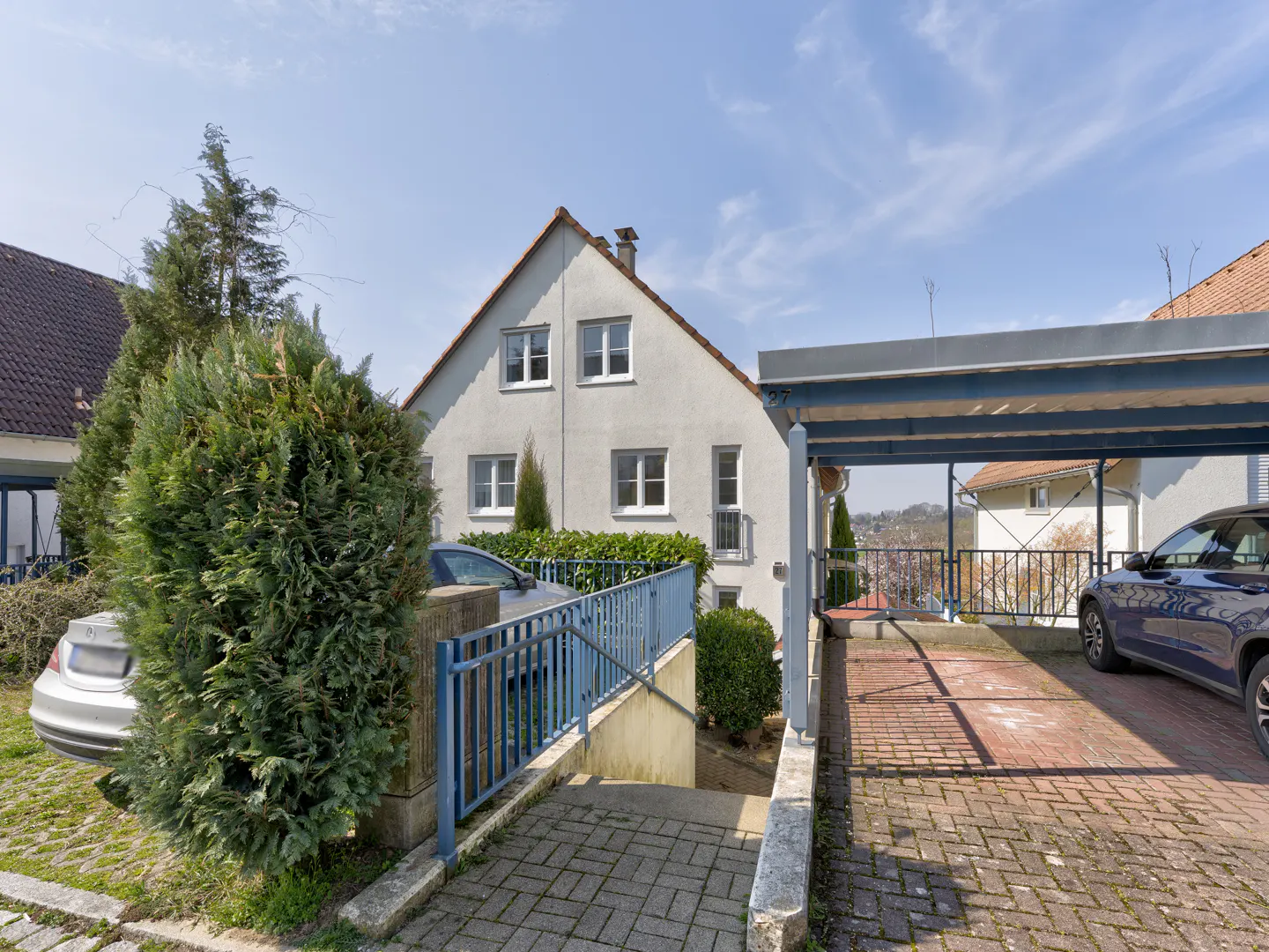 Two-story gray house with a carport and blue railings, cars parked in the driveway.