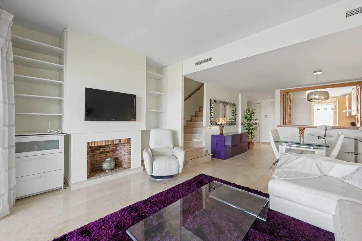 Bright living room with white walls, a purple rug, and a glass coffee table. A TV is mounted above a brick fireplace. A staircase is visible in the background.