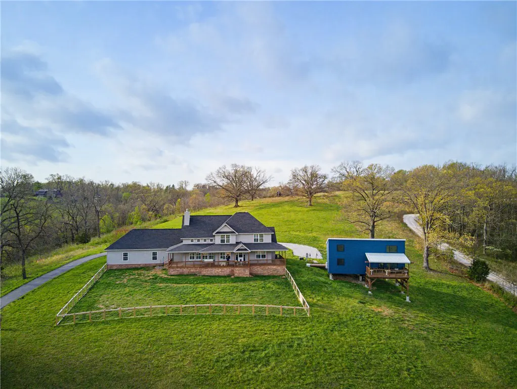 Aerial view of a gray two-story house with a porch, a blue outbuilding, and a green lawn on a cloudy day.