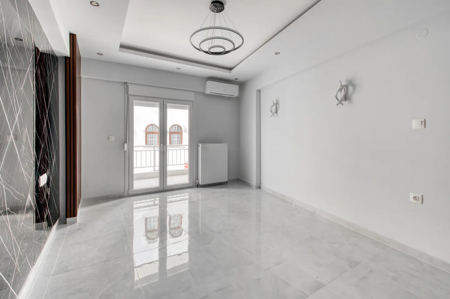 Bright, empty room with gray marble floors, white walls, and a modern chandelier. Balcony doors offer a view of a building across the street.