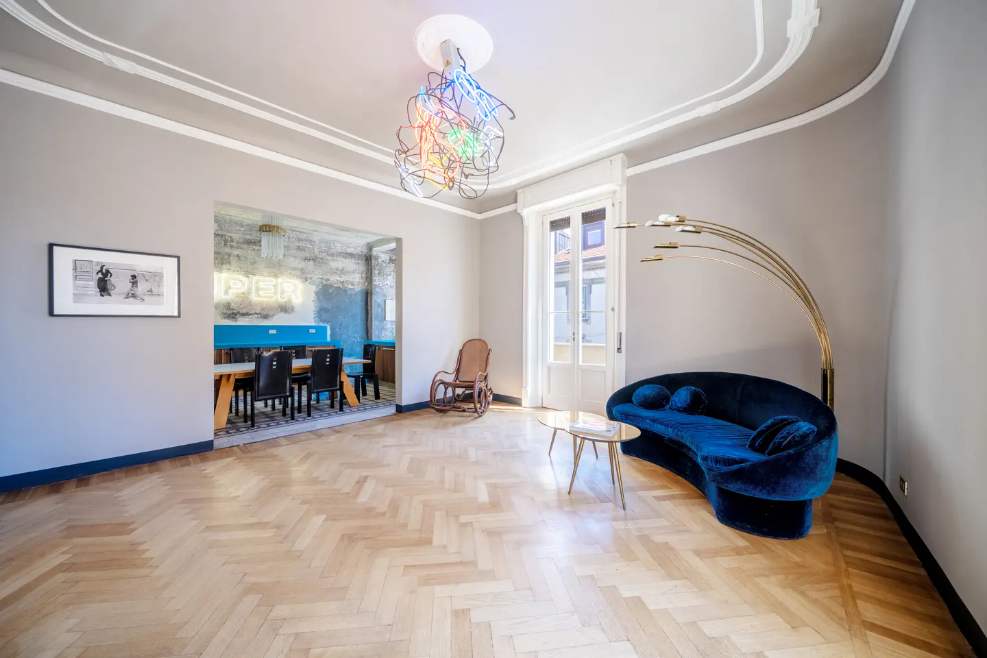 A living room with herringbone wood floors, a blue velvet sofa, and a colorful chandelier. A dining room is visible through an archway.